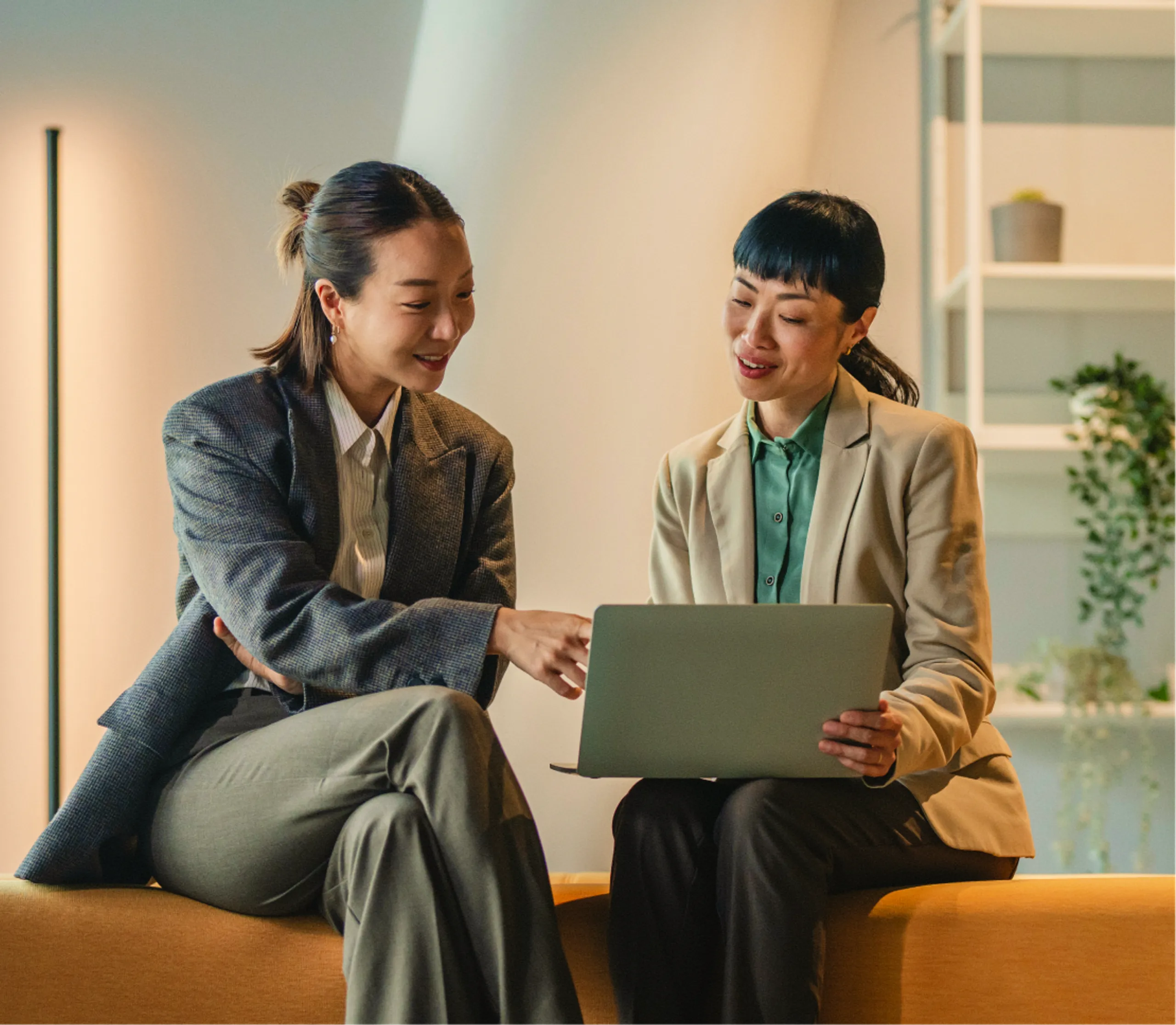 Two women in business attire sit on a couch, smiling and looking at a laptop together in a modern office setting with plants and shelves in the background.