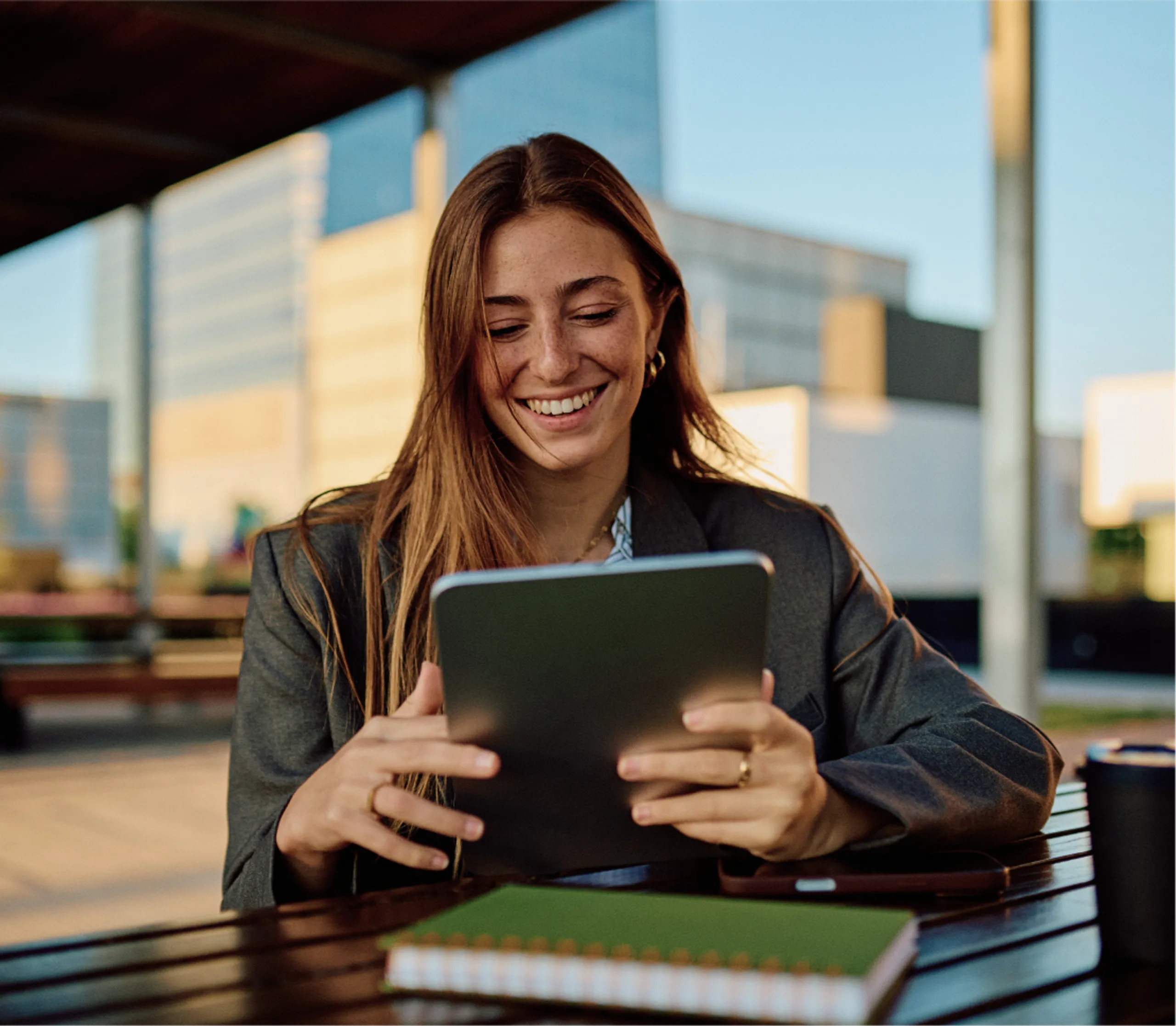 A woman with long brown hair smiles while using a tablet outdoors at a table. She is wearing a blazer, and a closed green notebook lies in front of her. Modern buildings are visible in the background.