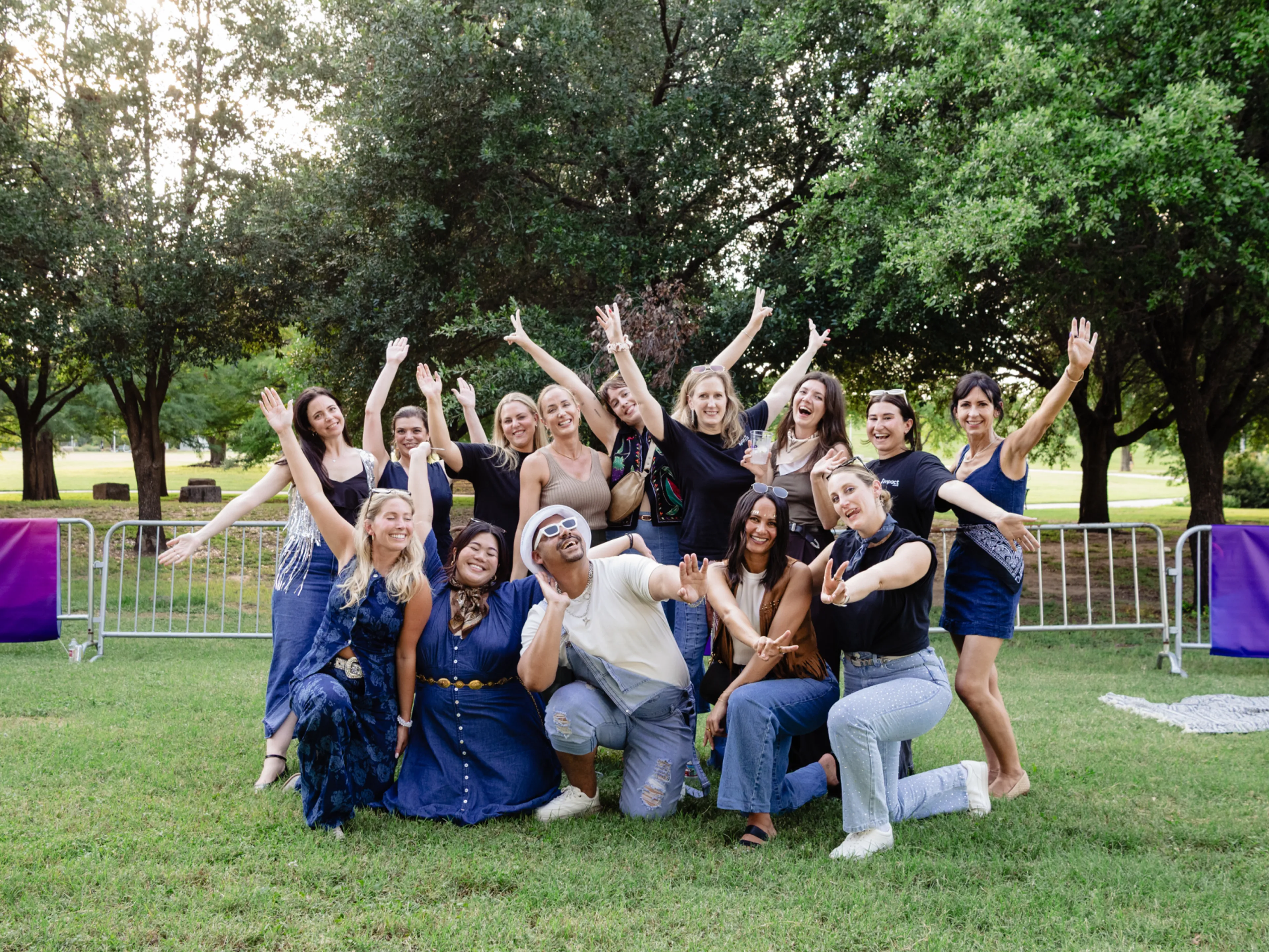 A group of people pose outdoors on grass, smiling and raising their arms enthusiastically. Trees and a metal fence are visible in the background under a bright sky.