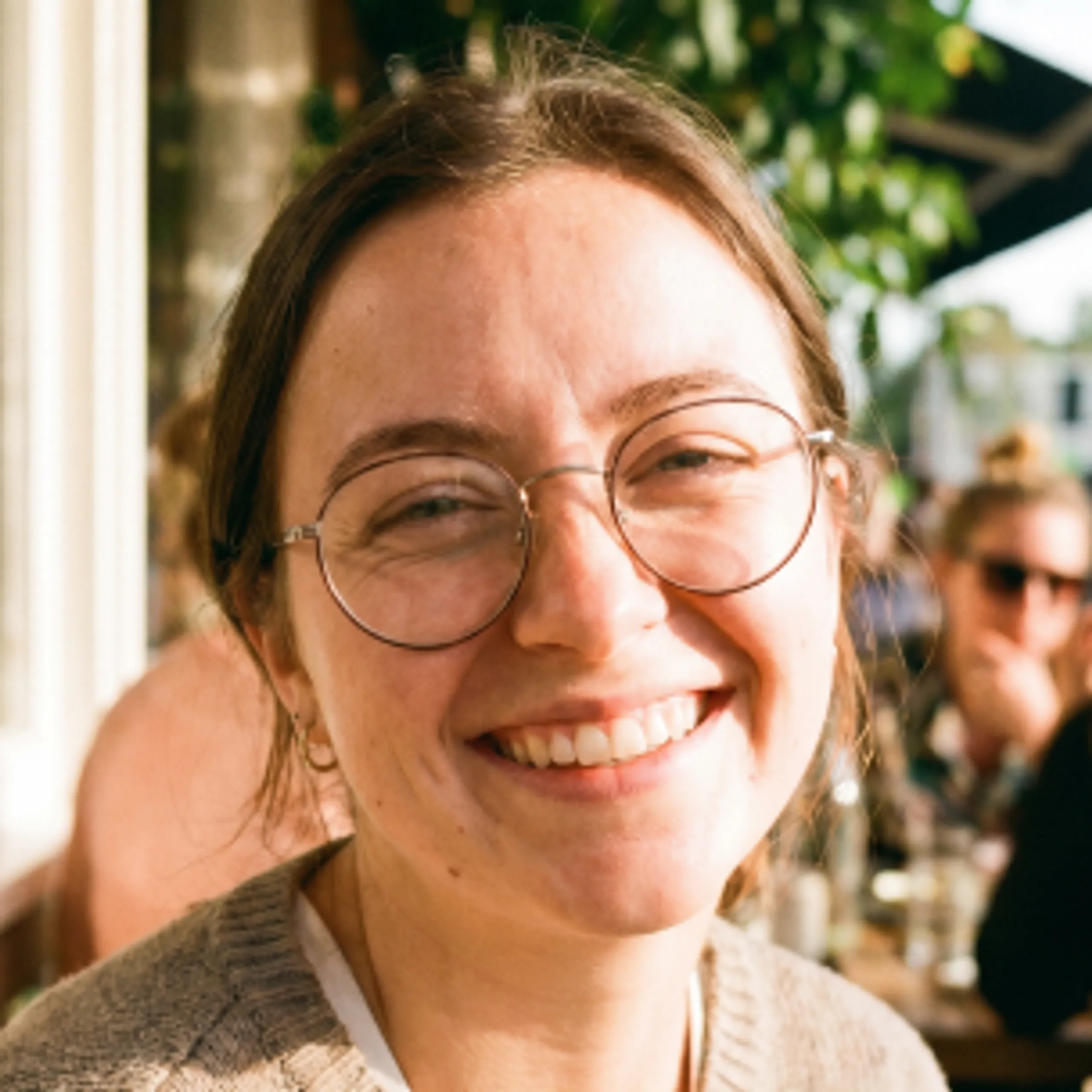 A person with glasses and brown hair smiles warmly while sitting outdoors at a sunlit café, with other people and greenery in the blurred background.