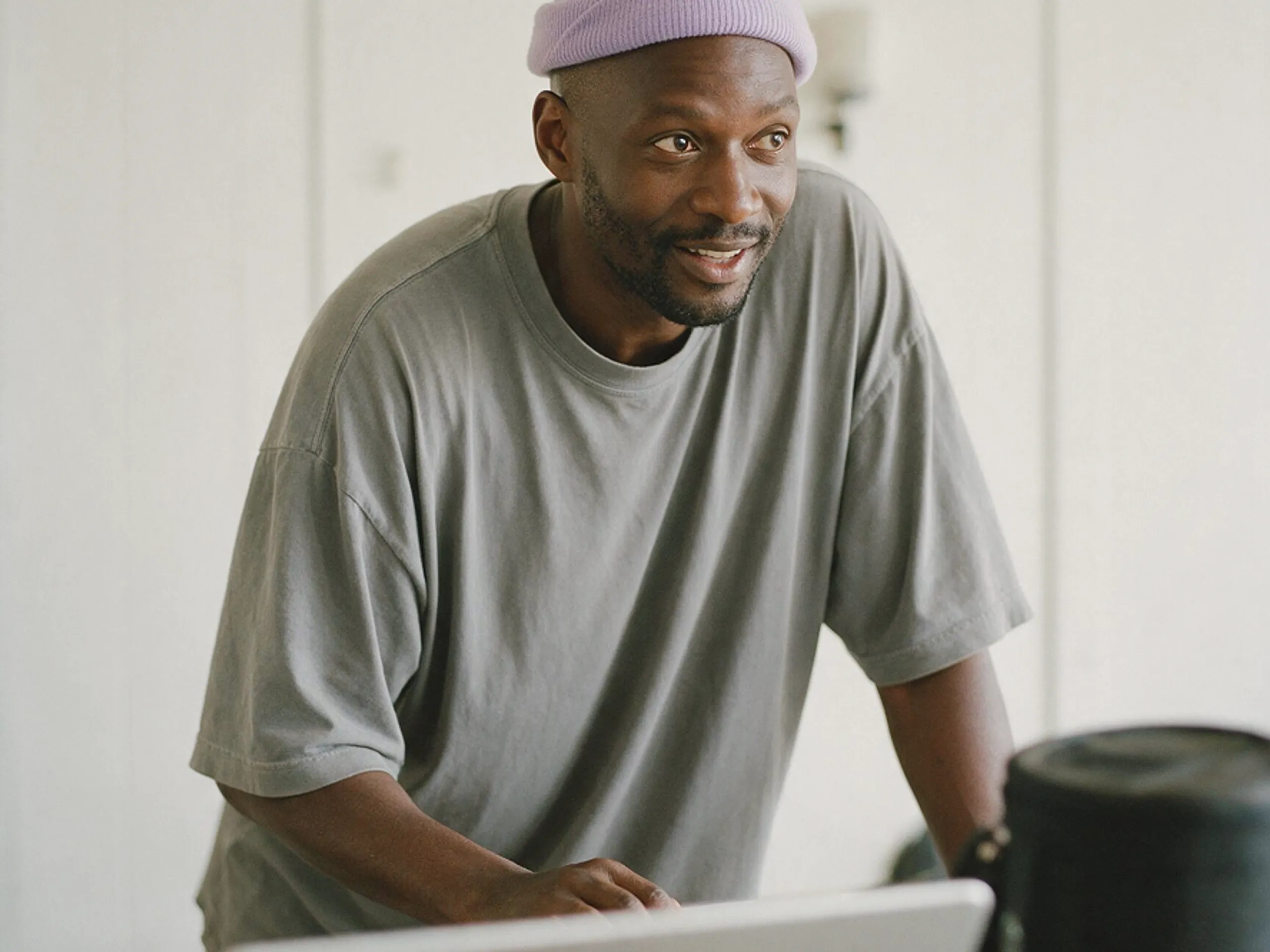A man wearing a purple beanie and gray t-shirt leans forward and smiles, looking toward a laptop in a bright room.
