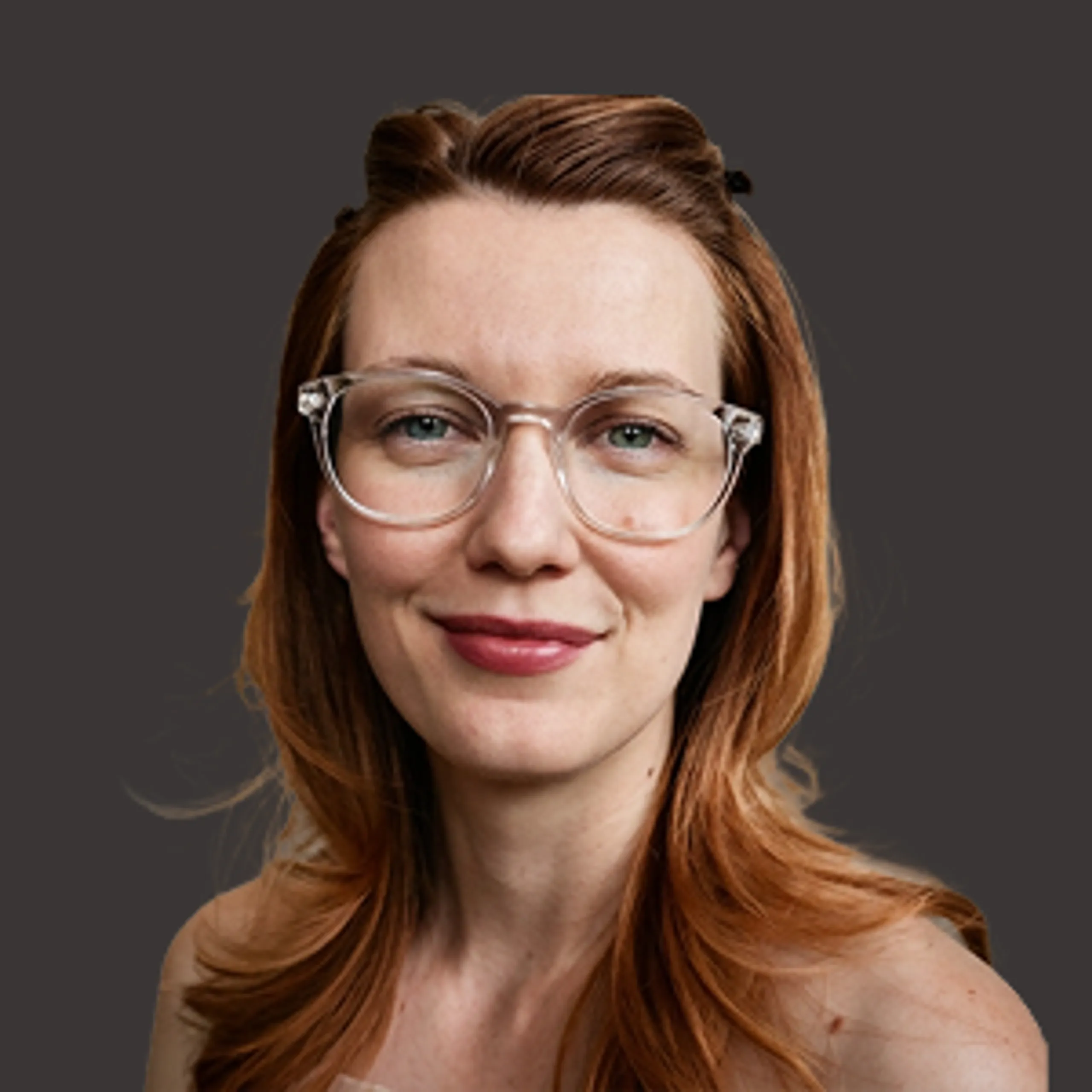 A woman with long reddish-brown hair, wearing clear-framed glasses, smiles slightly while looking at the camera. The background is a solid dark gray.