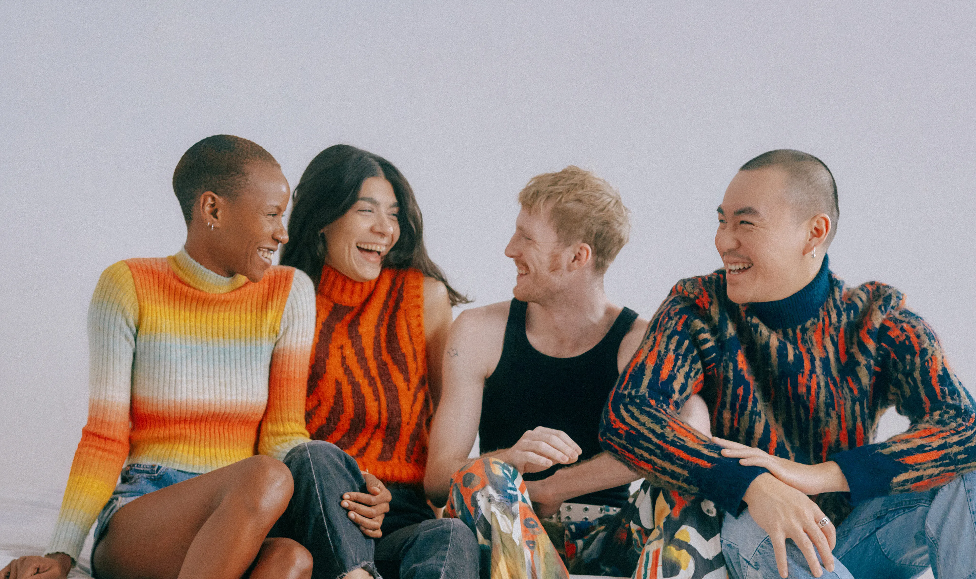 Four people sit side by side, smiling and laughing together. They wear colorful, patterned sweaters and casual pants. The background is plain and light-colored, creating a cheerful, relaxed atmosphere.