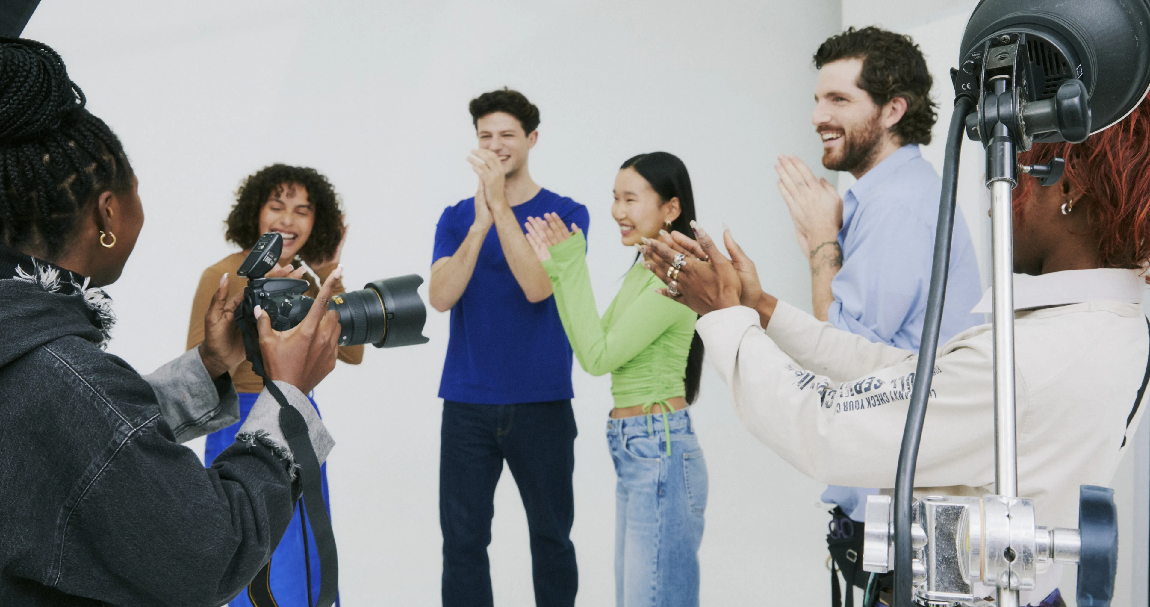 A group of four people smile and clap while facing a photographer in a studio with lighting equipment. The scene looks cheerful and energetic.