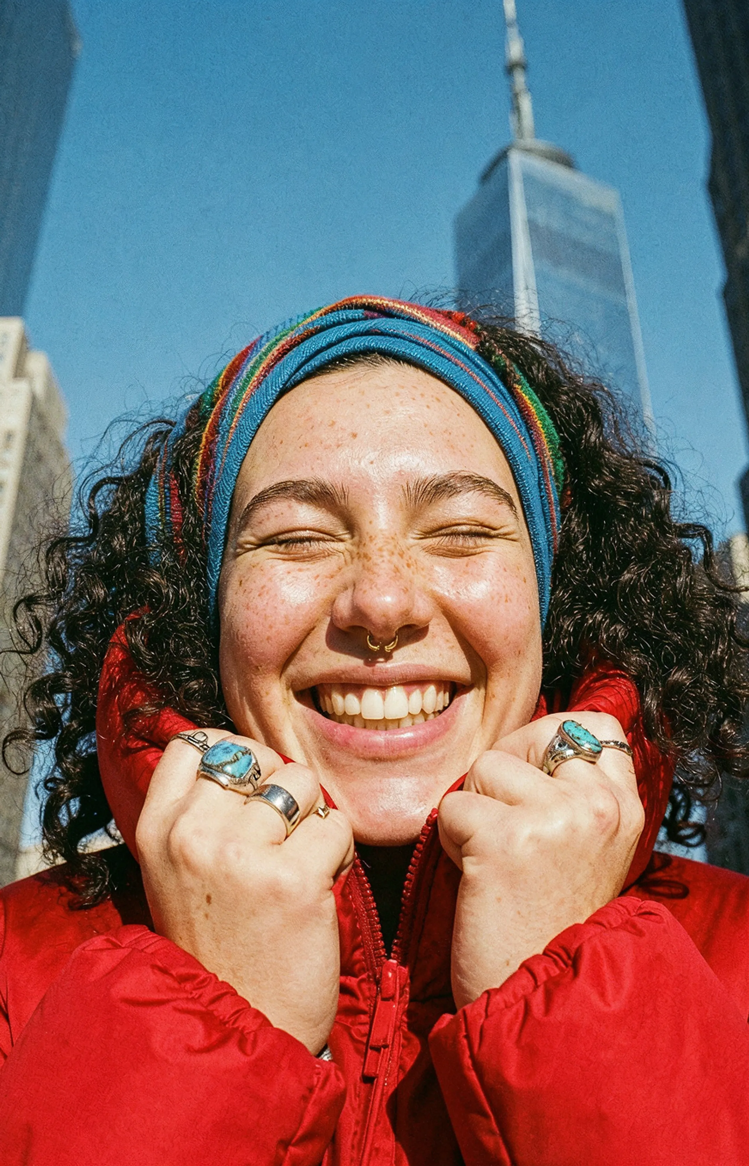 A person with curly hair and a colorful headband smiles widely, eyes closed, holding their red jacket collar. Skyscrapers and a blue sky are in the background.