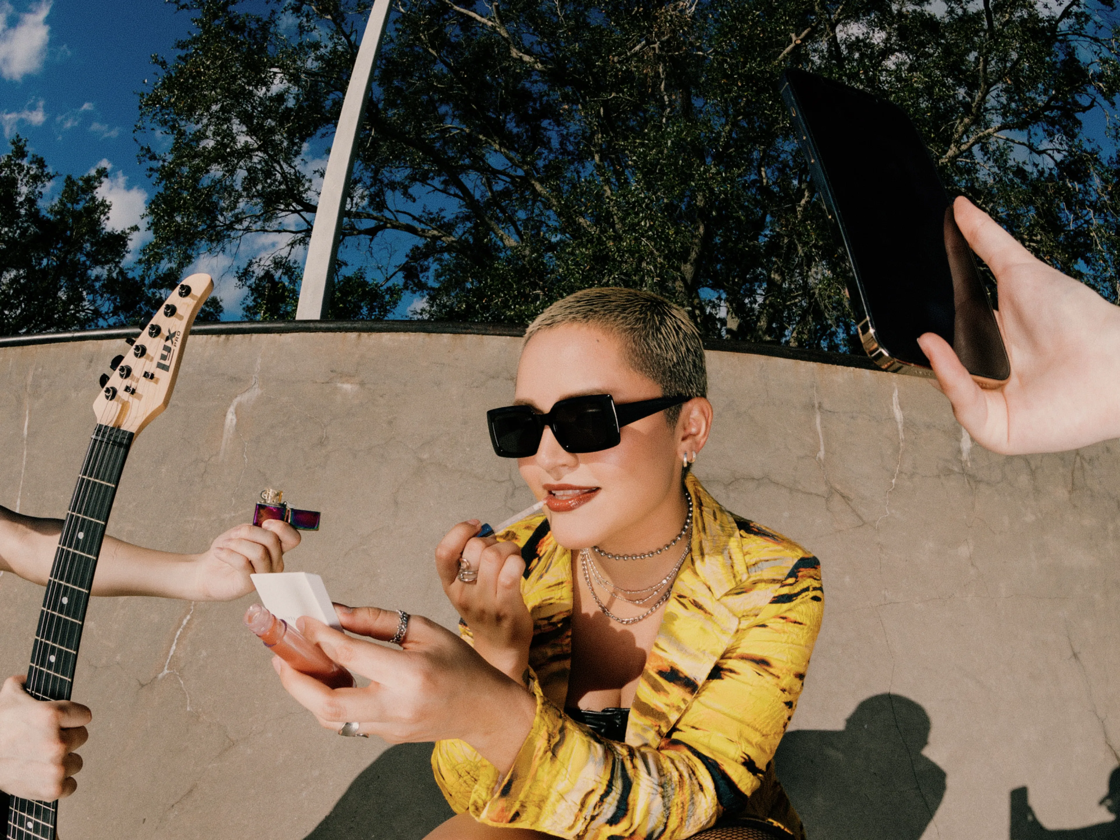 A person with short hair and sunglasses applies lipstick, wearing a yellow outfit. Hands offer a lighter, a guitar, and a tablet. Trees and blue sky are in the background.