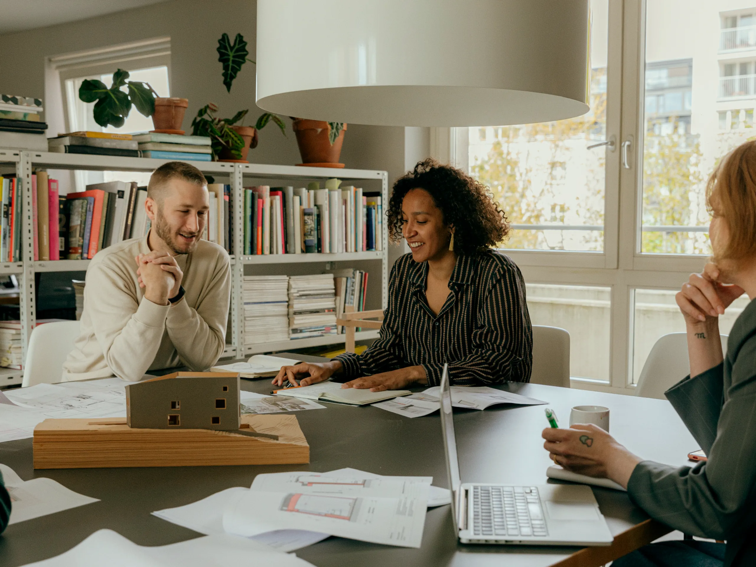 Three people sit around a table with papers, a laptop, and a building model, discussing ideas in a modern, bright office with bookshelves and plants in the background.
