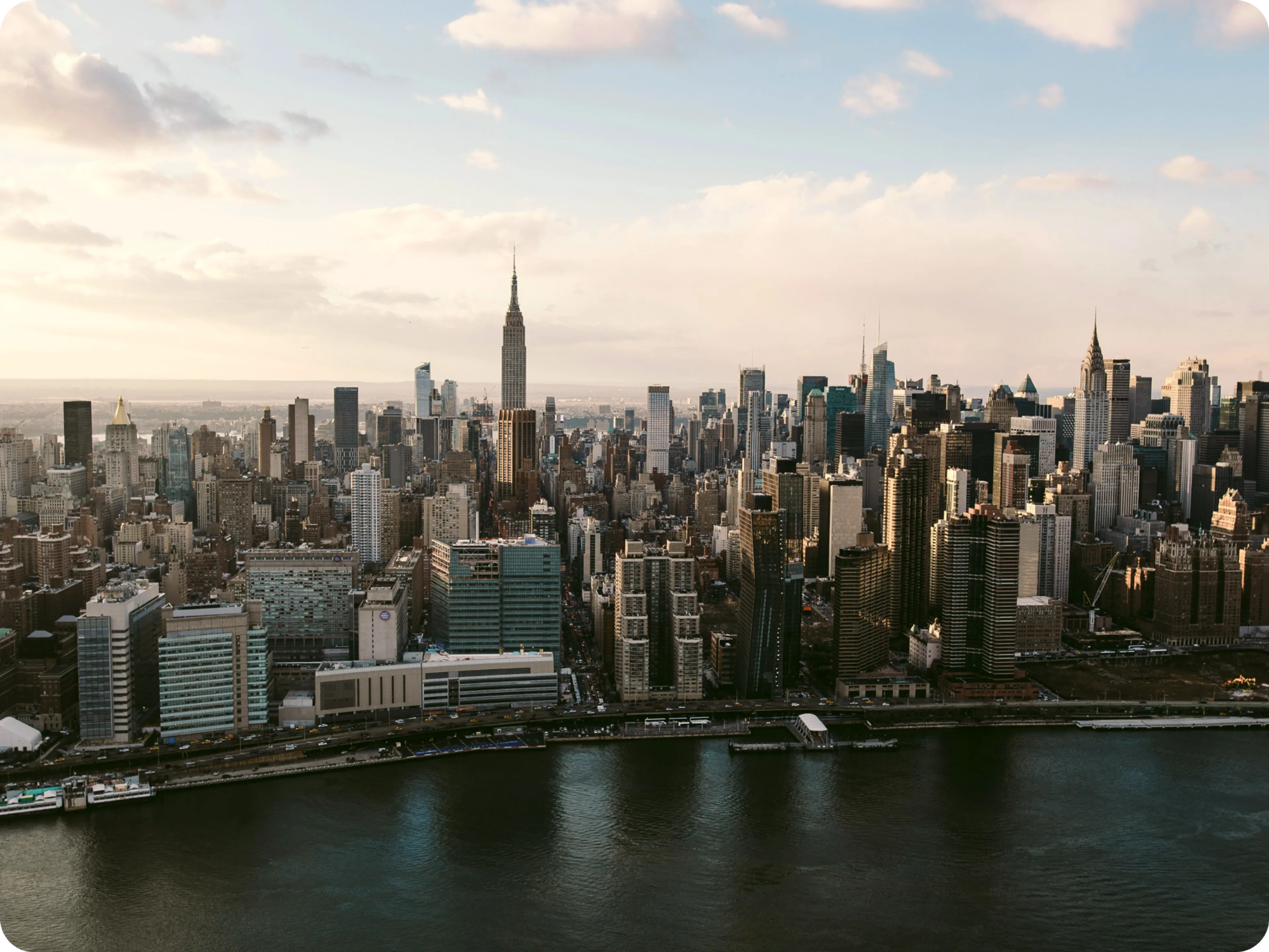 Aerial view of New York City skyline with the Empire State Building centered, skyscrapers surrounding it, and the East River in the foreground under a partly cloudy sky.