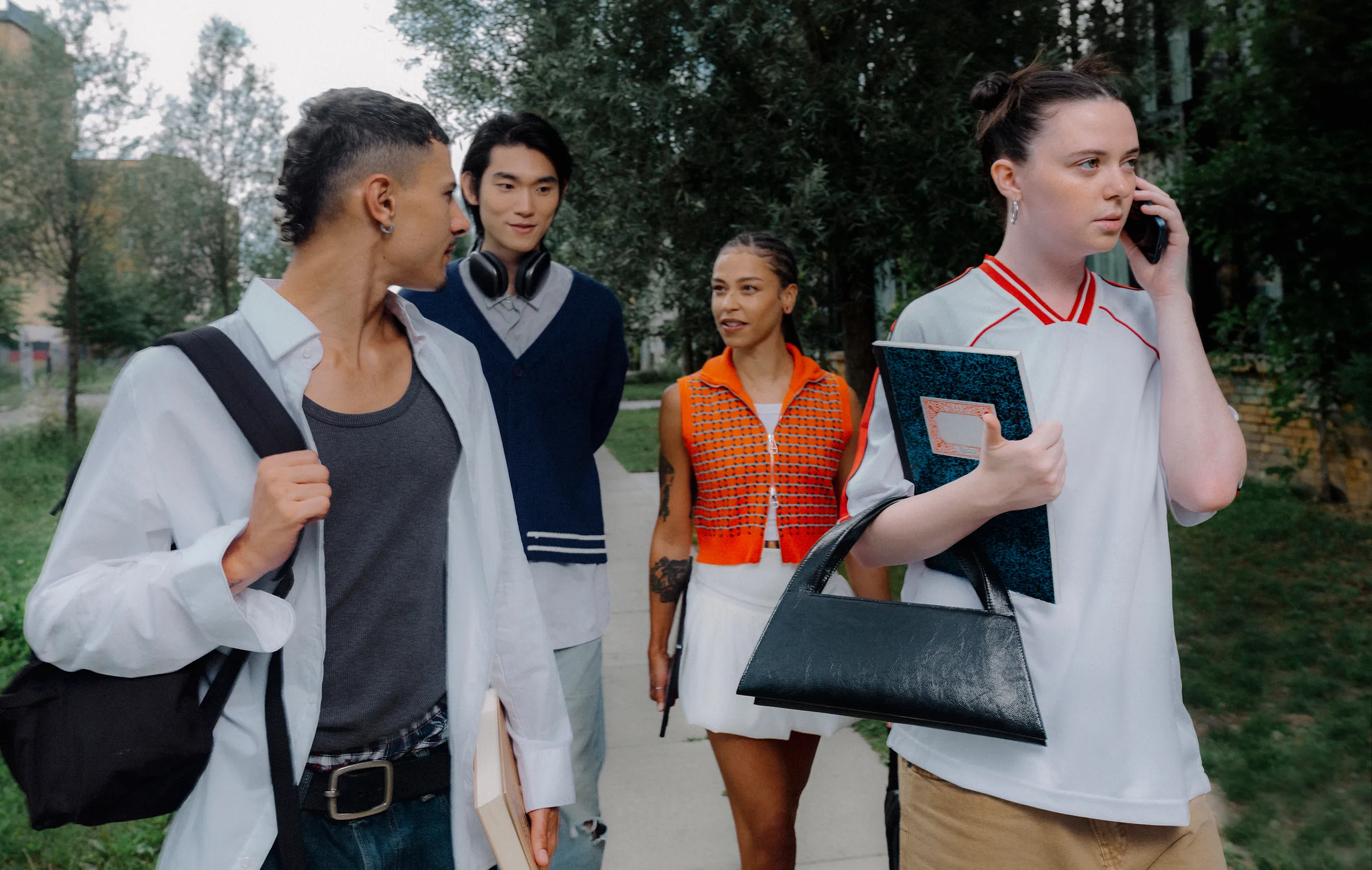 Four young adults walk outdoors. One holds a phone to their ear, while others chat and carry bags or notebooks. Trees and buildings are in the background.