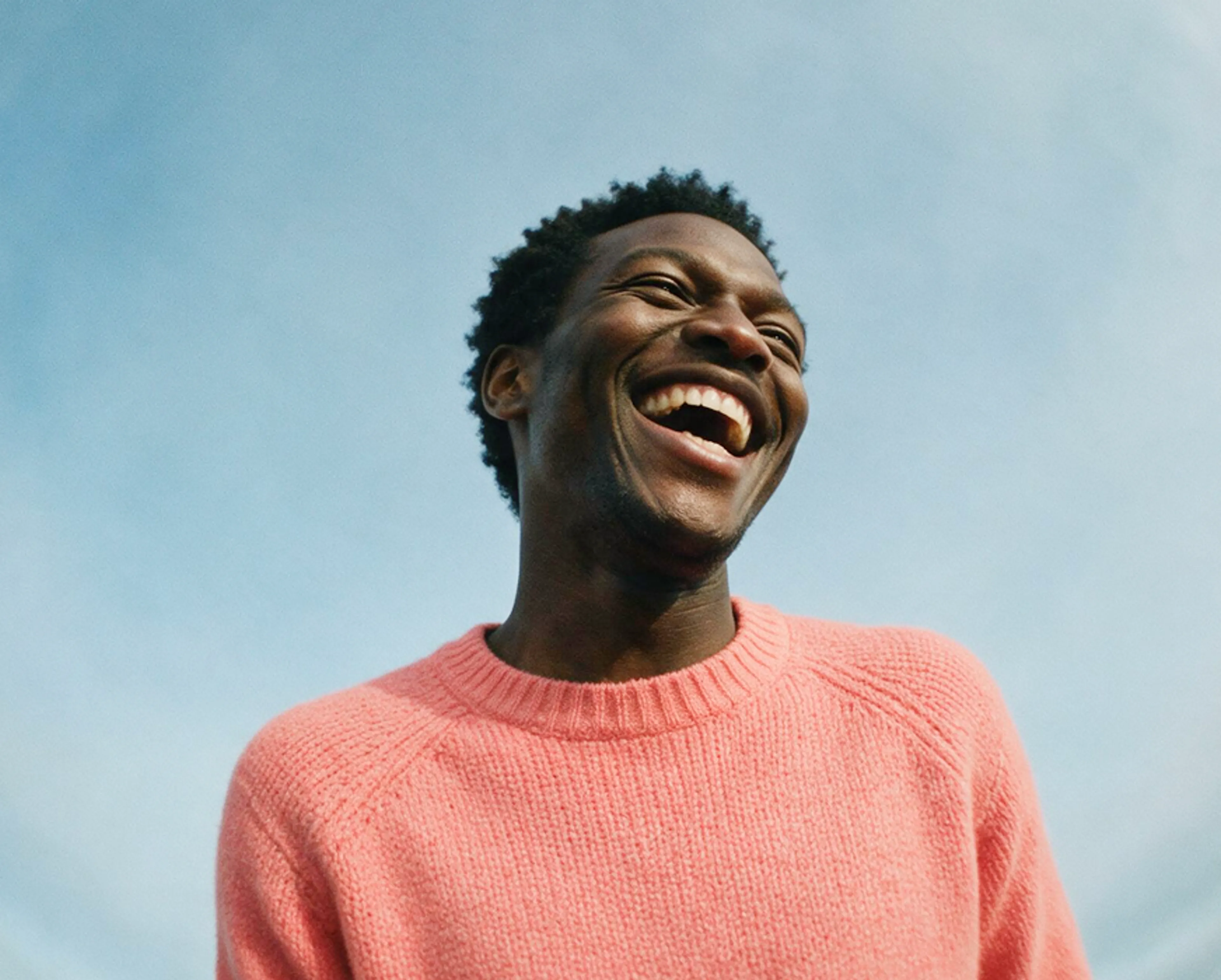 A person wearing a pink sweater smiles widely and laughs, looking up against a clear blue sky.