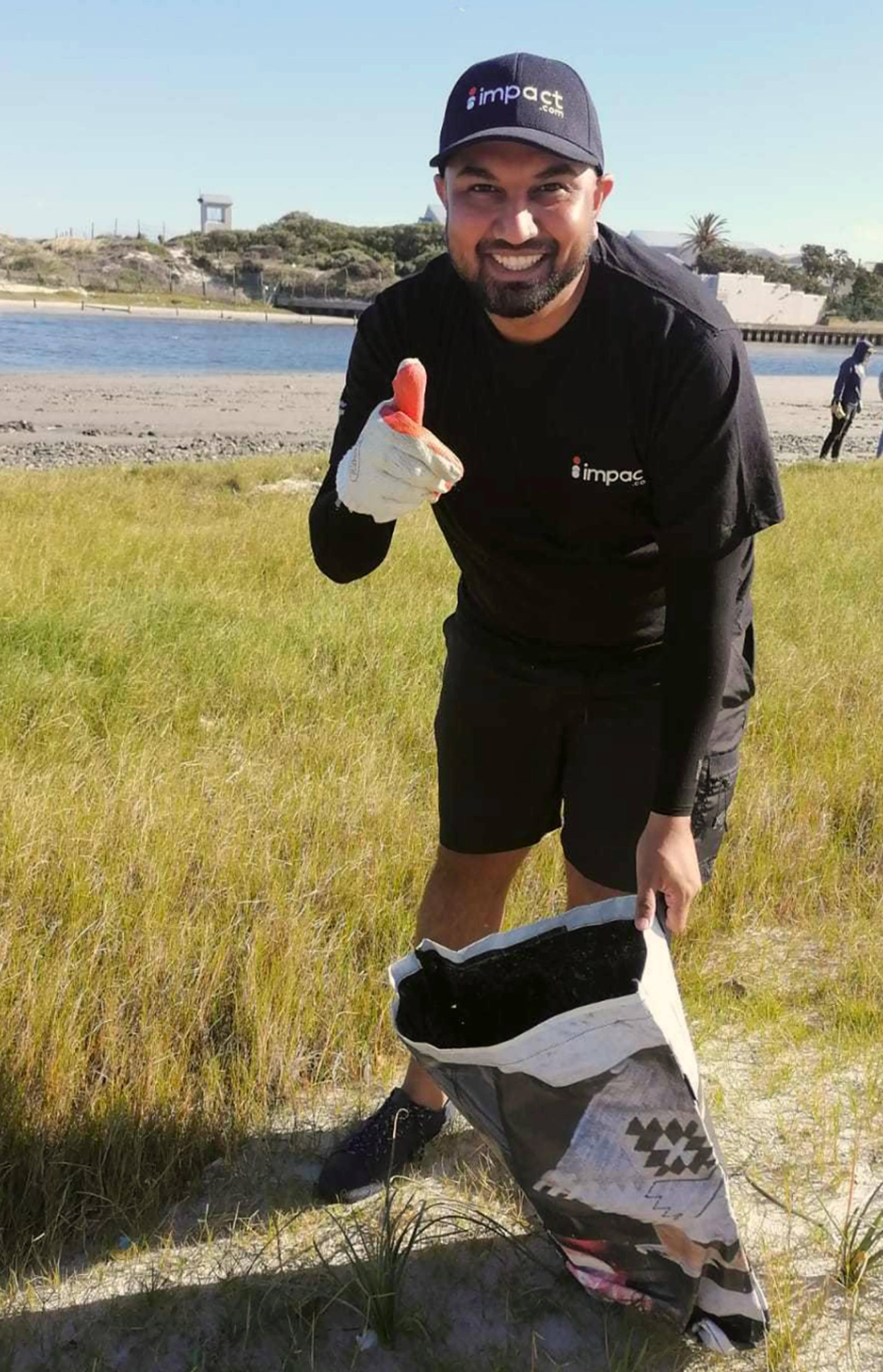 A man wearing gloves and an impact shirt picks up litter on a grassy beach, smiling and holding a piece of trash, with a bag and water in the background.