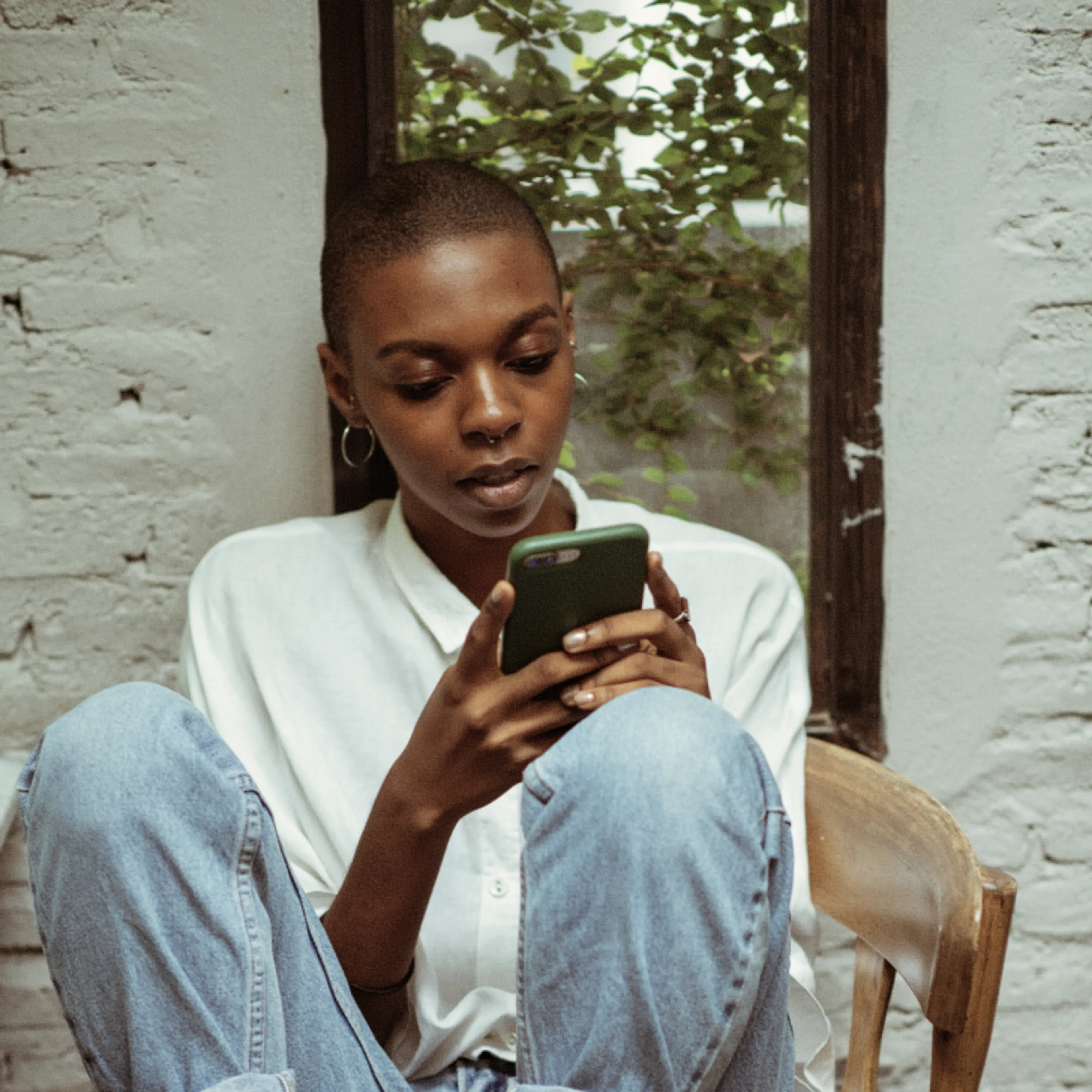 A person with short hair, wearing a white shirt and jeans, sits on a chair looking at a smartphone. There is a large mirror behind them reflecting green plants and a white brick wall.