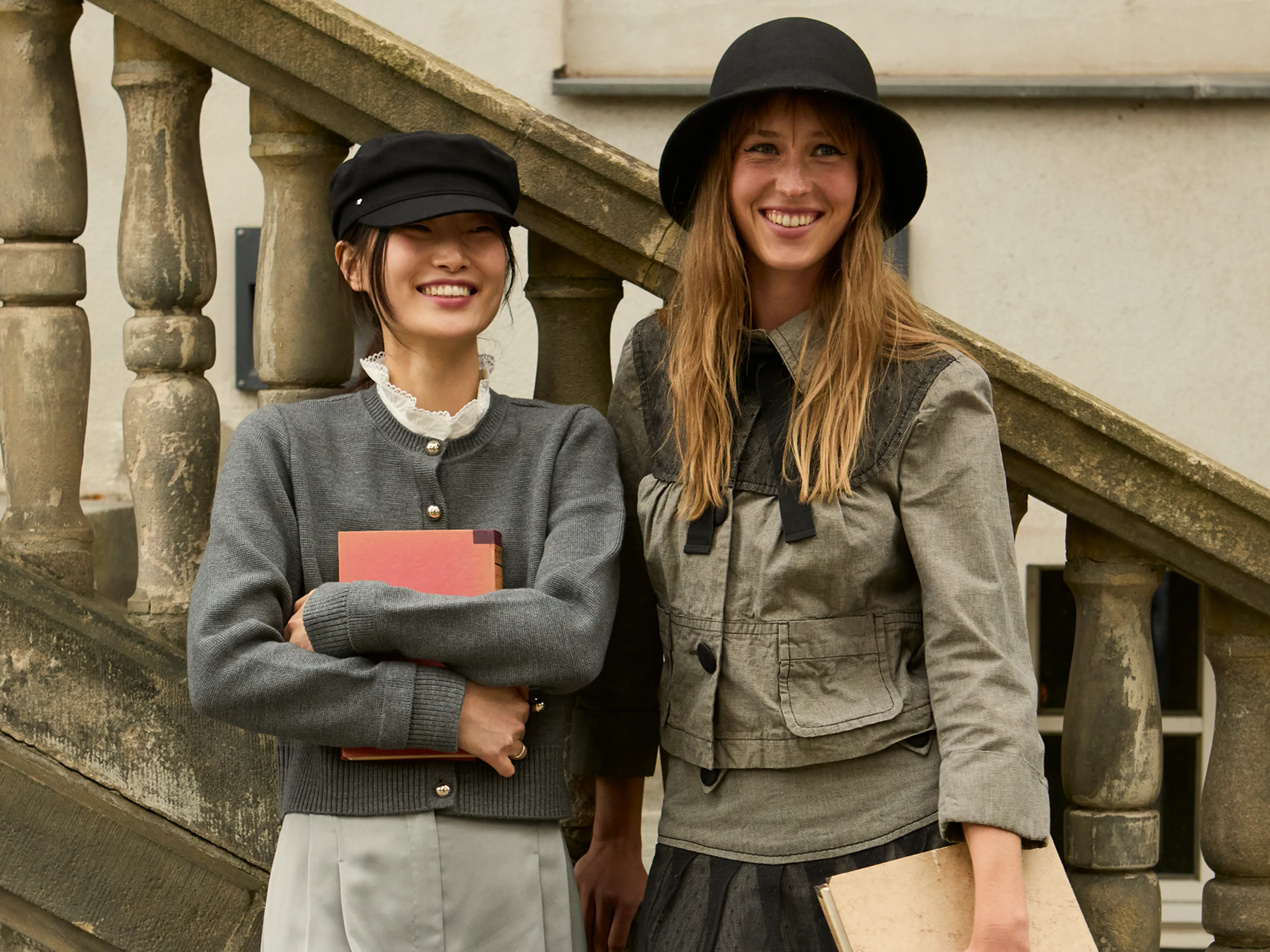 Two women stand outside by stone stairs, smiling. One wears a black cap and gray sweater, holding red books. The other has a black hat, green jacket, and holds a beige book. They appear happy and relaxed.