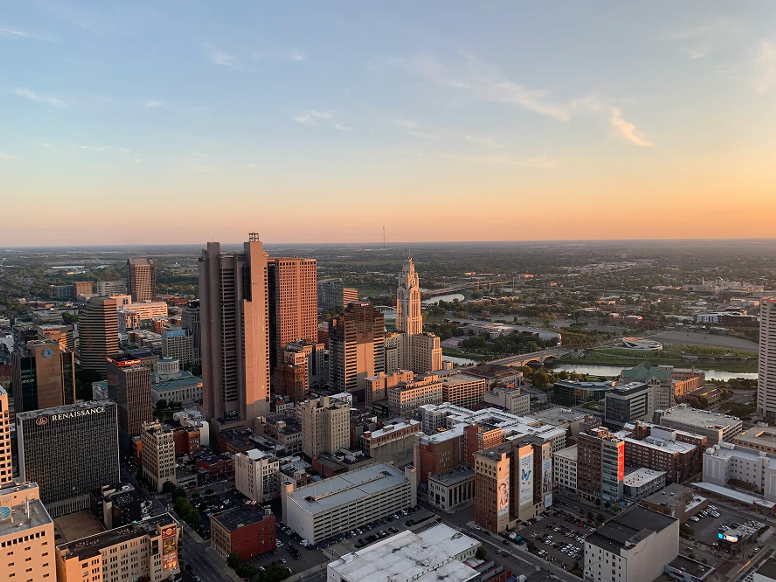 Aerial view of a city skyline at sunset, with tall buildings casting long shadows and a river visible in the background under a pastel sky.