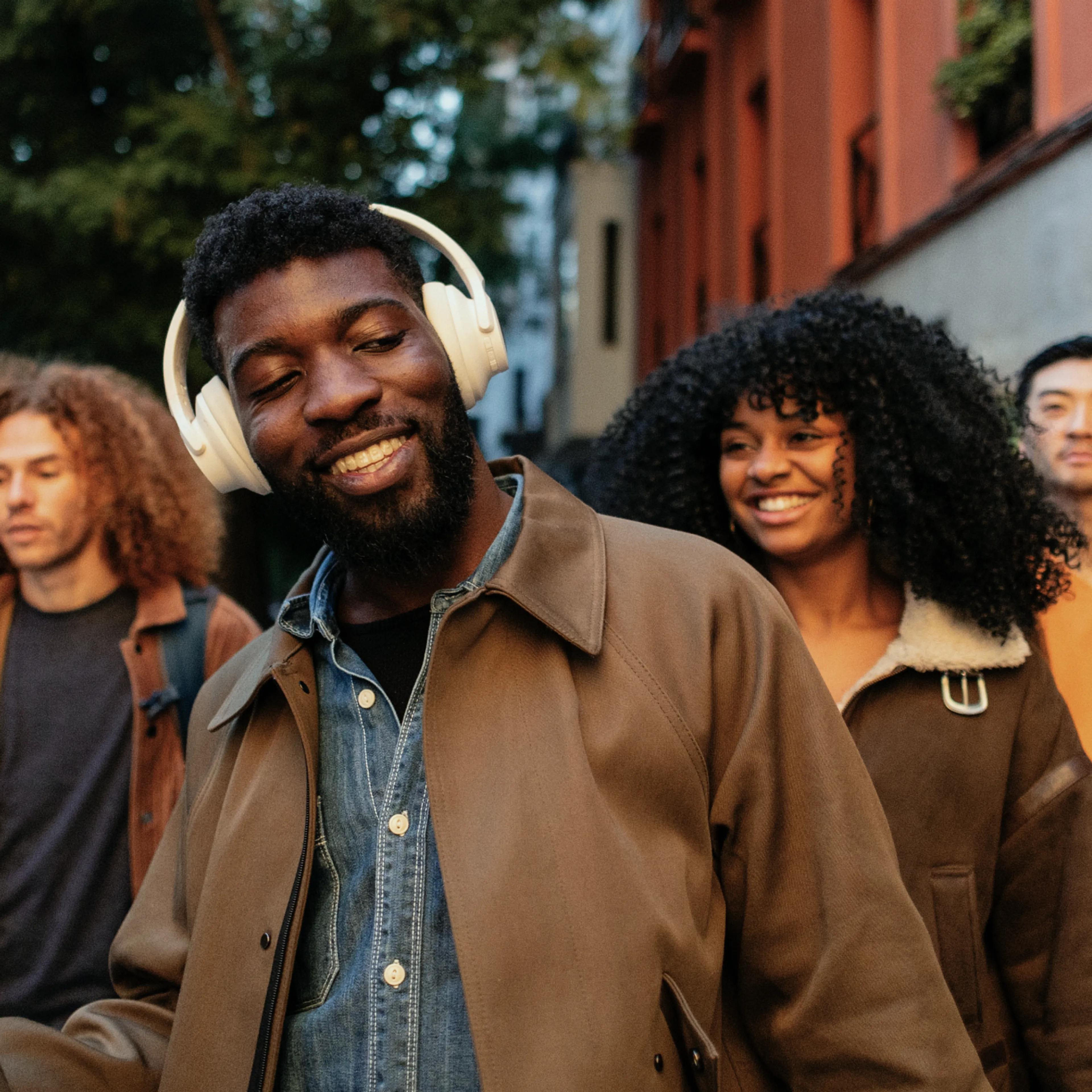 A man wearing headphones smiles while walking outdoors with friends. The group appears happy and relaxed, and trees and a red building are visible in the background.