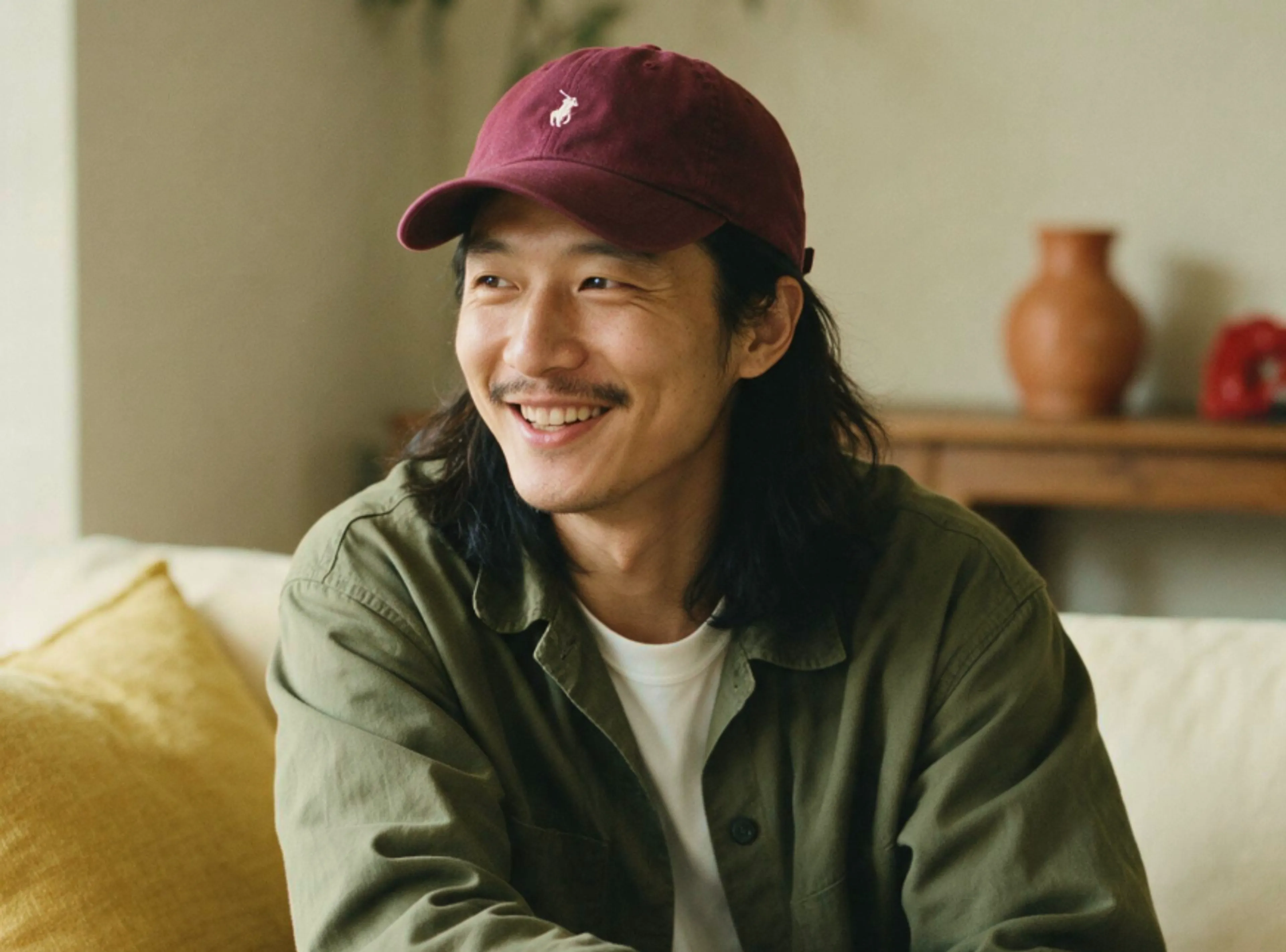 A person with long hair and a mustache, wearing a maroon cap, white shirt, and green jacket, sits on a couch smiling. A yellow pillow and wooden table with pottery are in the background.