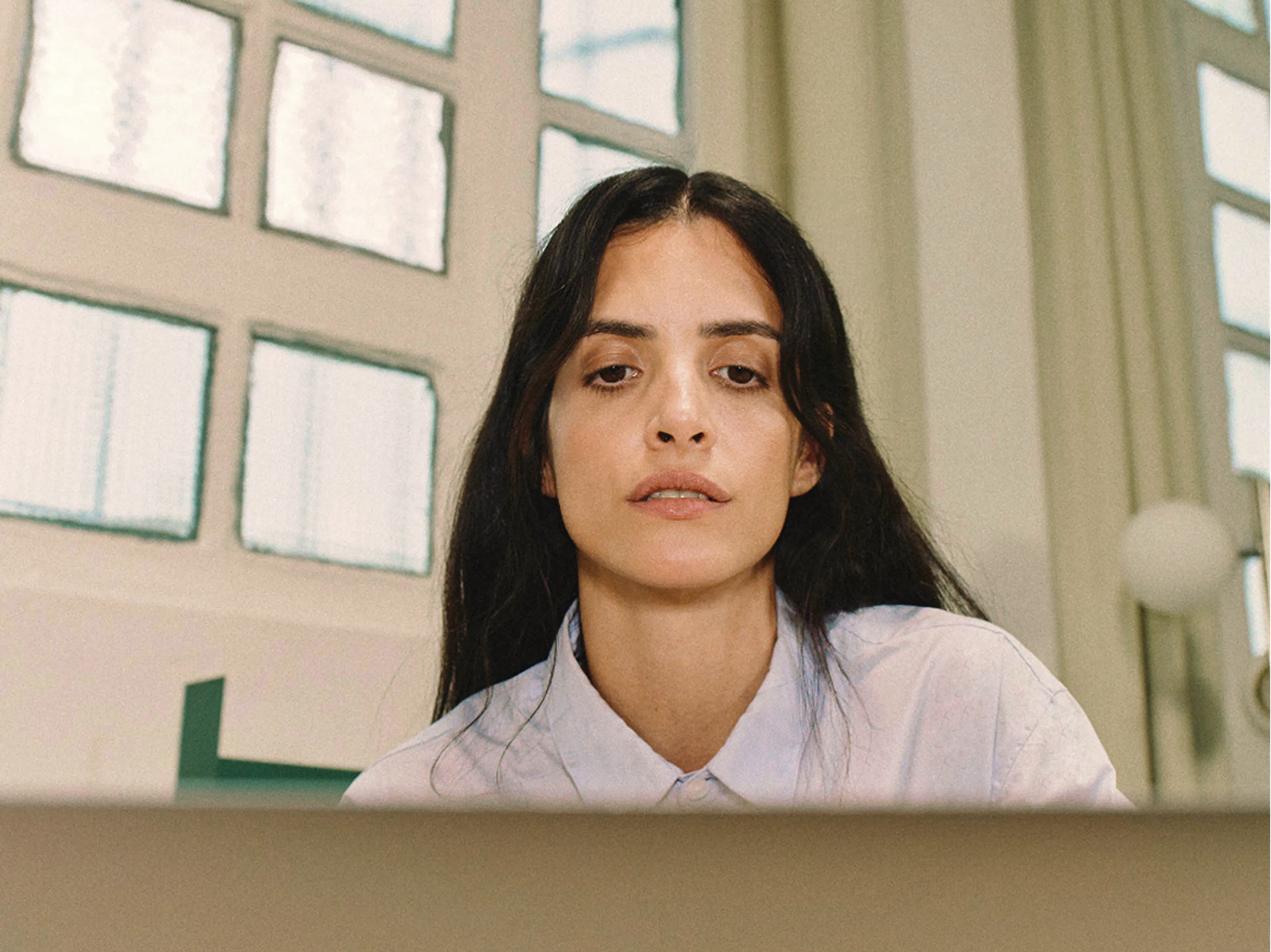 A woman with long dark hair, wearing a light blue shirt, looks at a laptop screen in a bright room with large windows and light curtains.