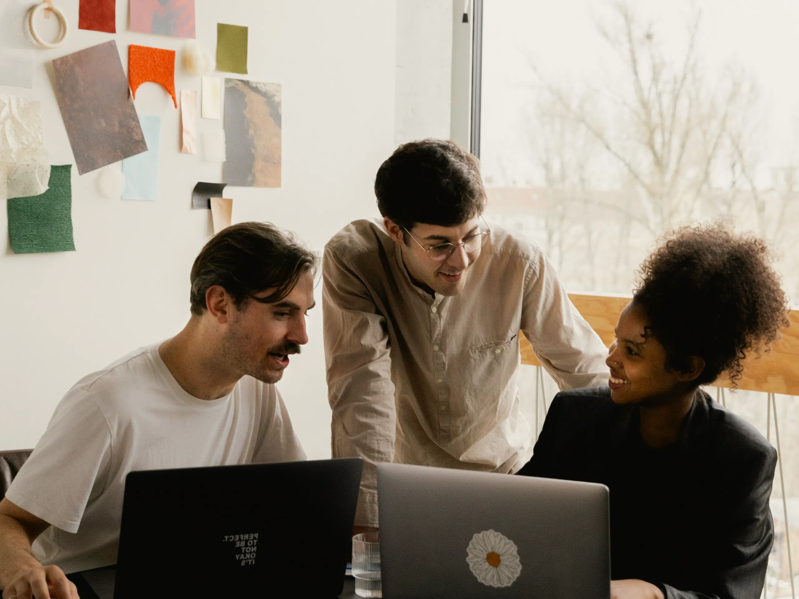 Three people sit and stand around a table with laptops, smiling and having a discussion. Artworks and color samples are pinned on the wall behind them, and a large window lets in natural light.