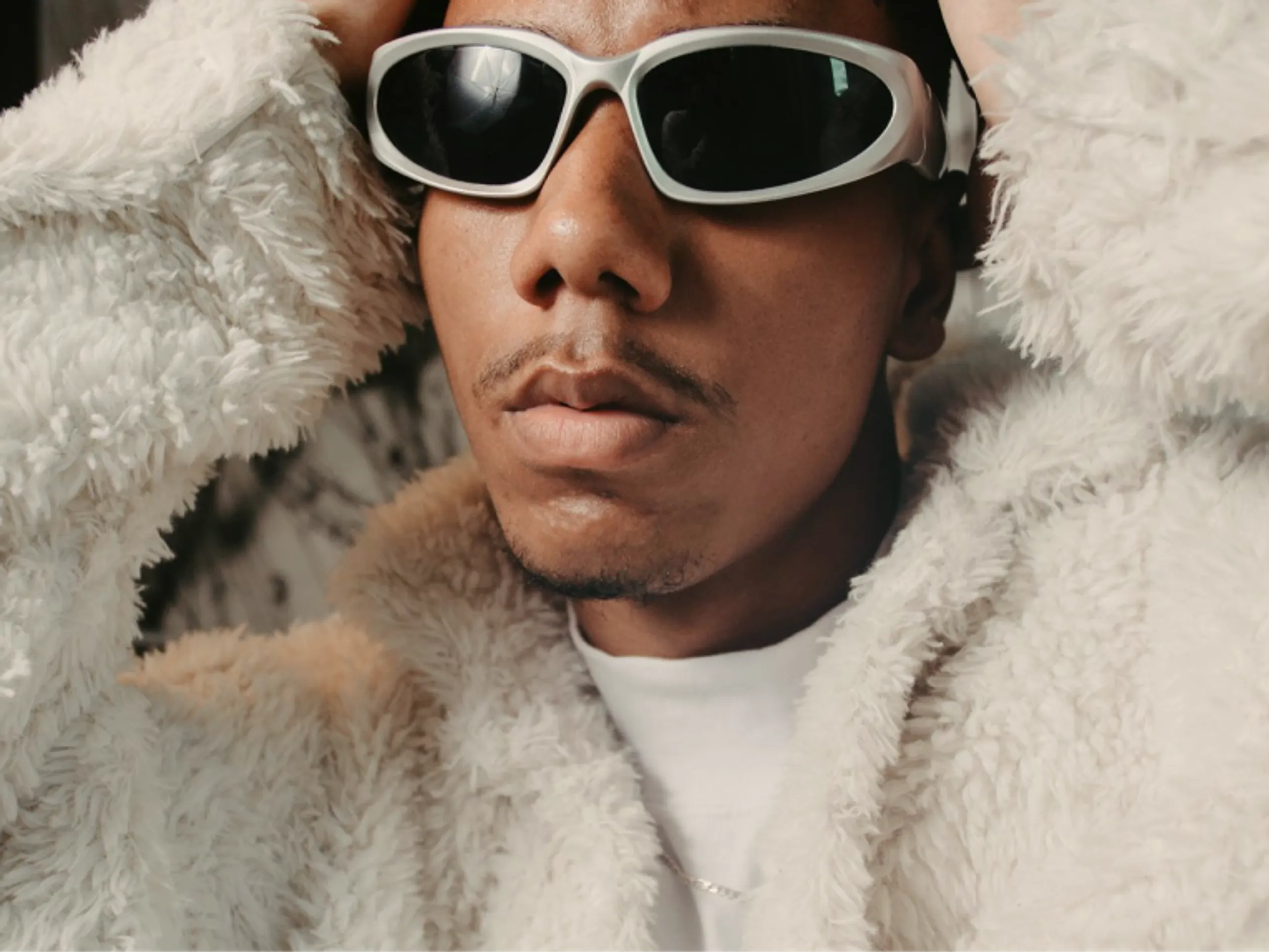 A person wearing white sunglasses and a fluffy white jacket looks into the camera, holding their hands near their head.