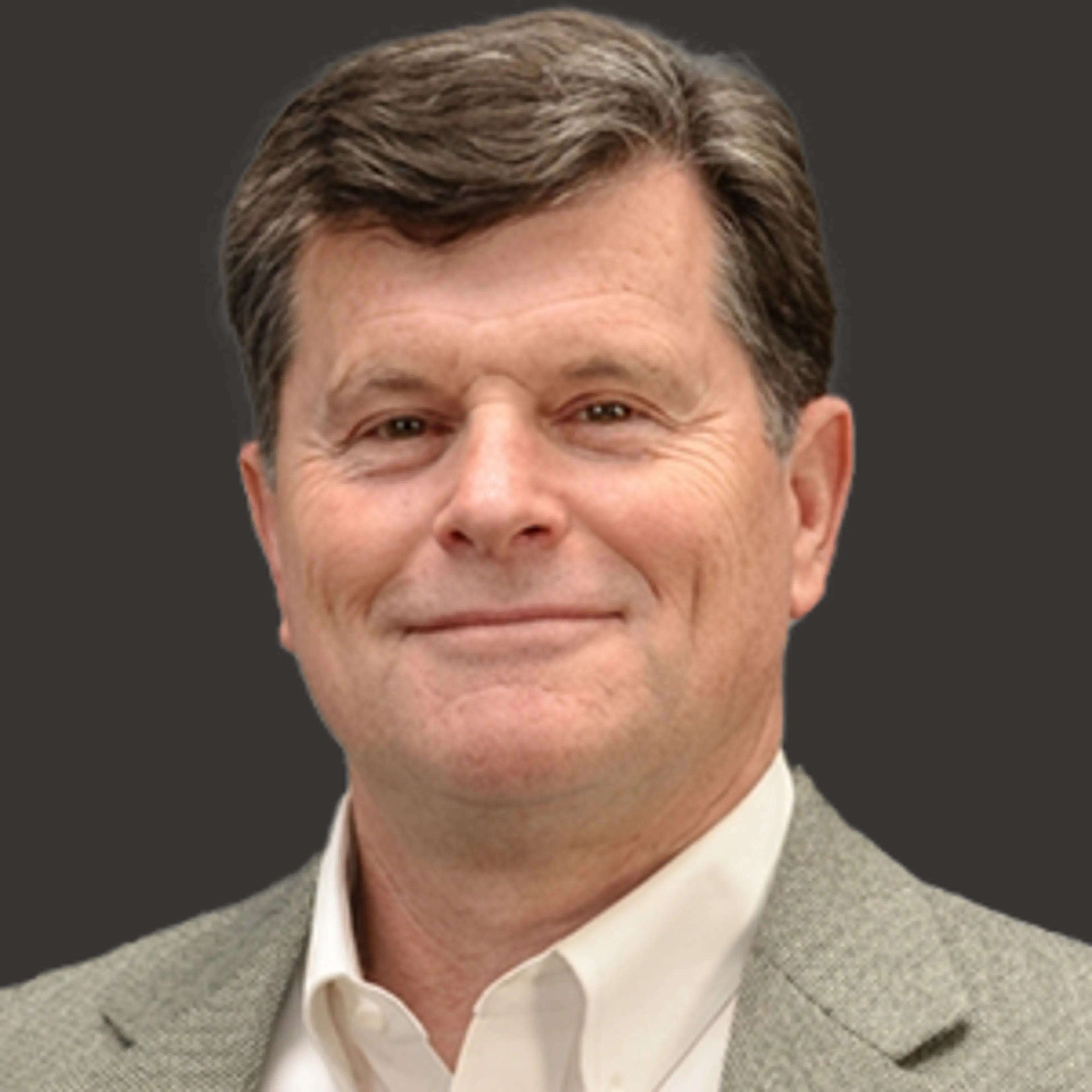A middle-aged man with short brown hair wearing a light gray blazer and white collared shirt, smiling in front of a plain dark background.