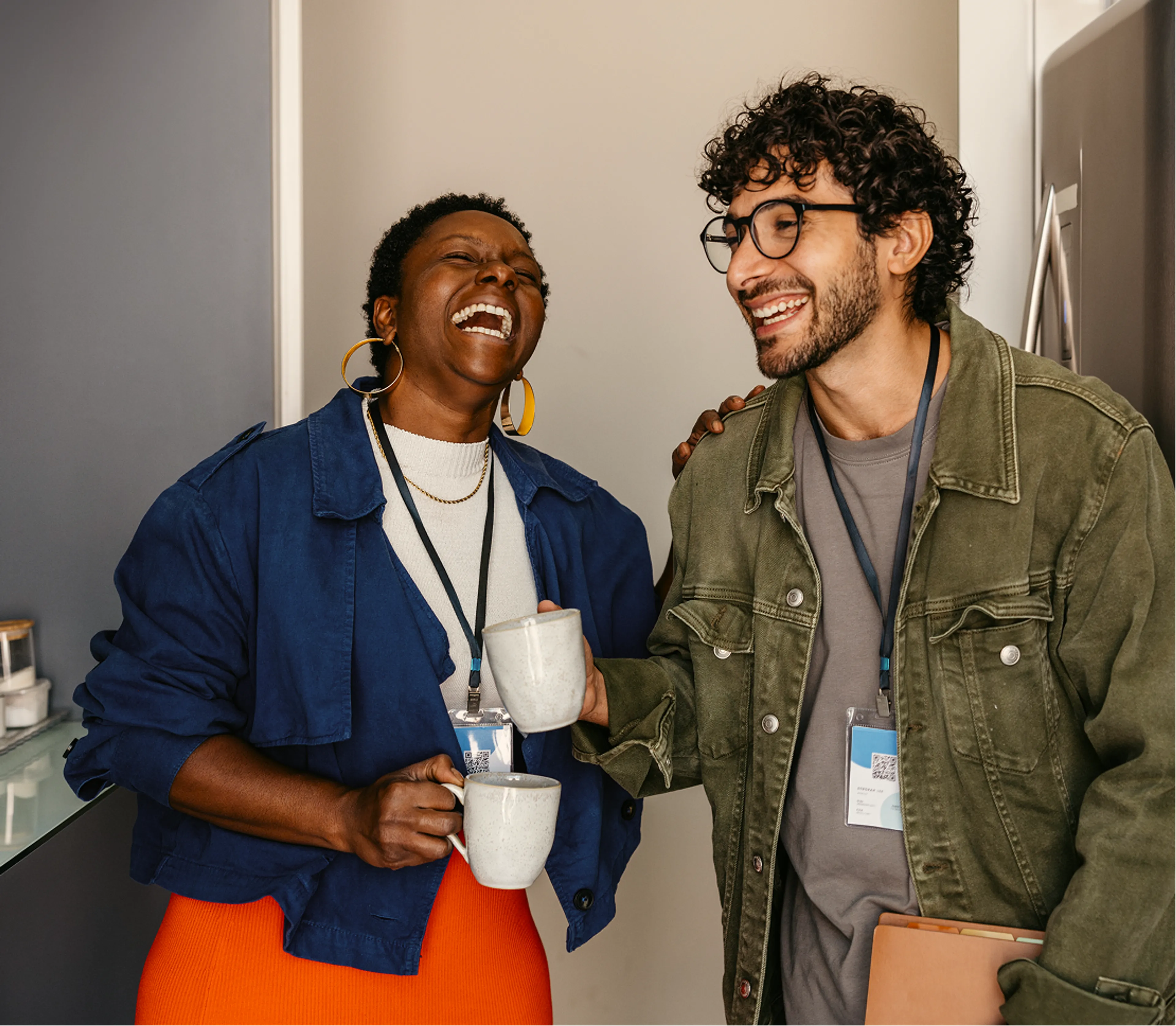 A Black woman in a blue jacket and a man with curly hair and glasses in an olive green jacket laugh together while holding mugs in an office breakroom, both wearing lanyards with identification badges.