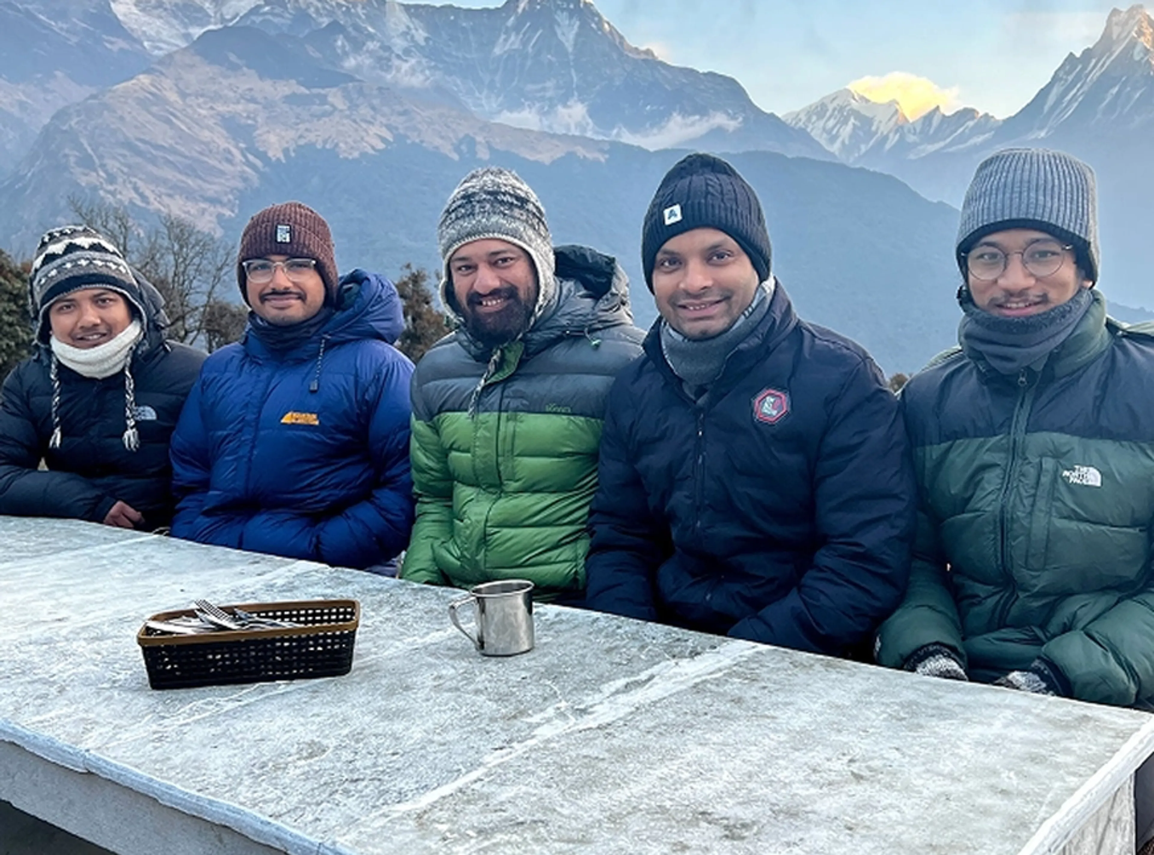 Five people in winter jackets and hats sit at a frosty outdoor table with a metal cup and basket, with snow-covered mountains in the background.