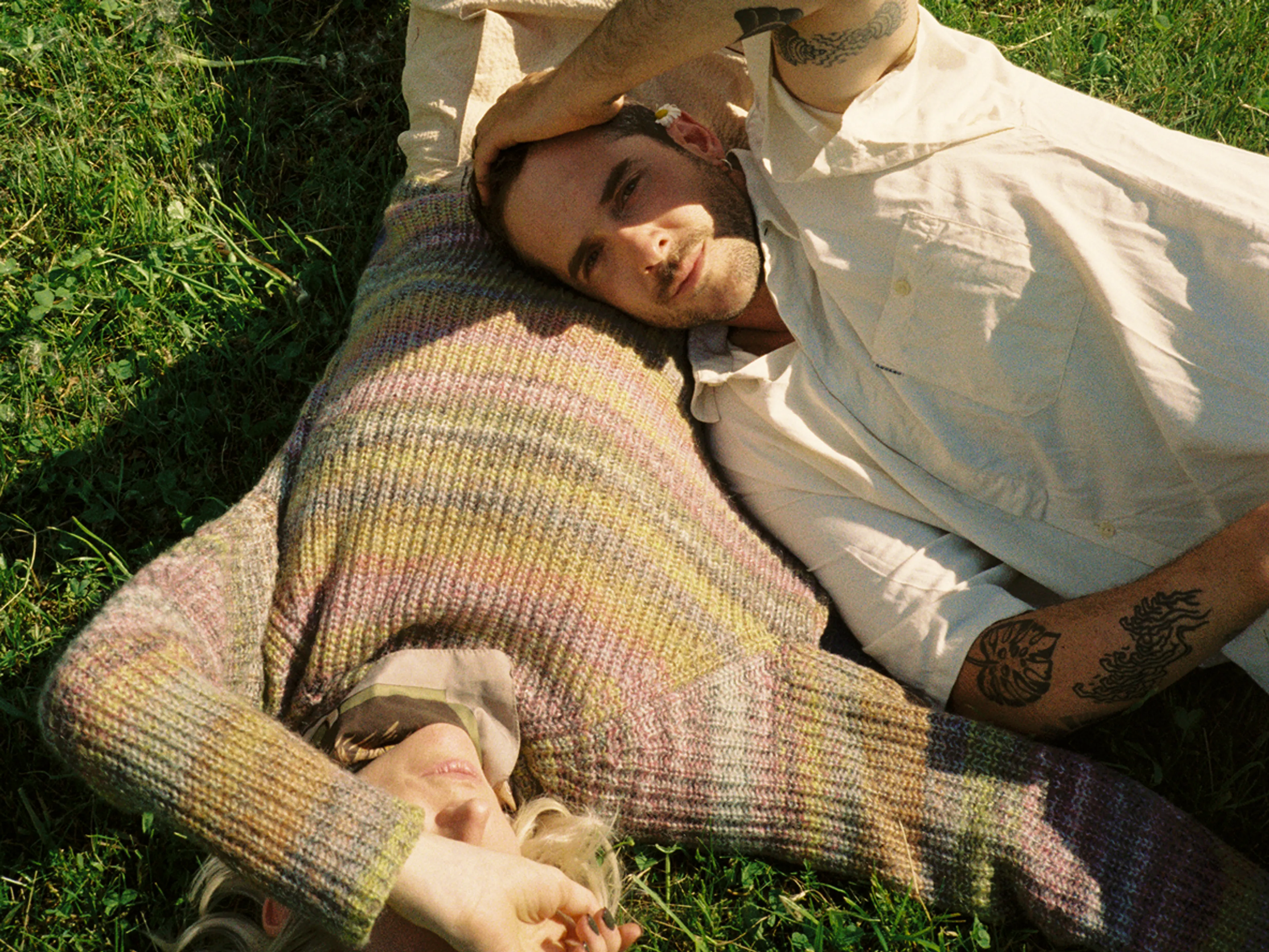 A couple lies on grass, heads close together. One person in a striped sweater covers their face, while the other, in a light shirt, looks at the camera with their hand gently resting on the others head.