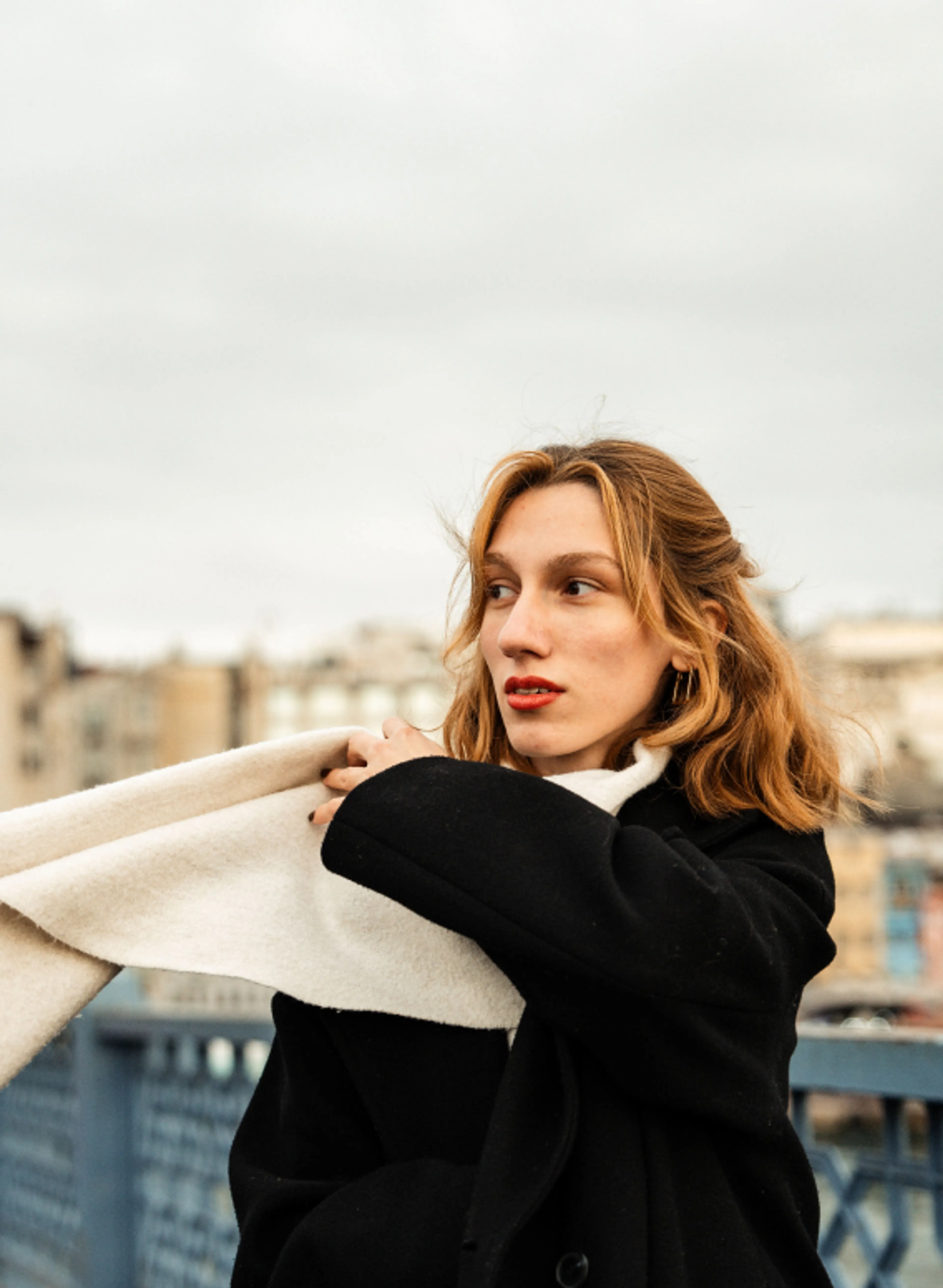 A woman with light brown hair wearing a black coat and white scarf stands outdoors on a cloudy day, with blurred buildings in the background.