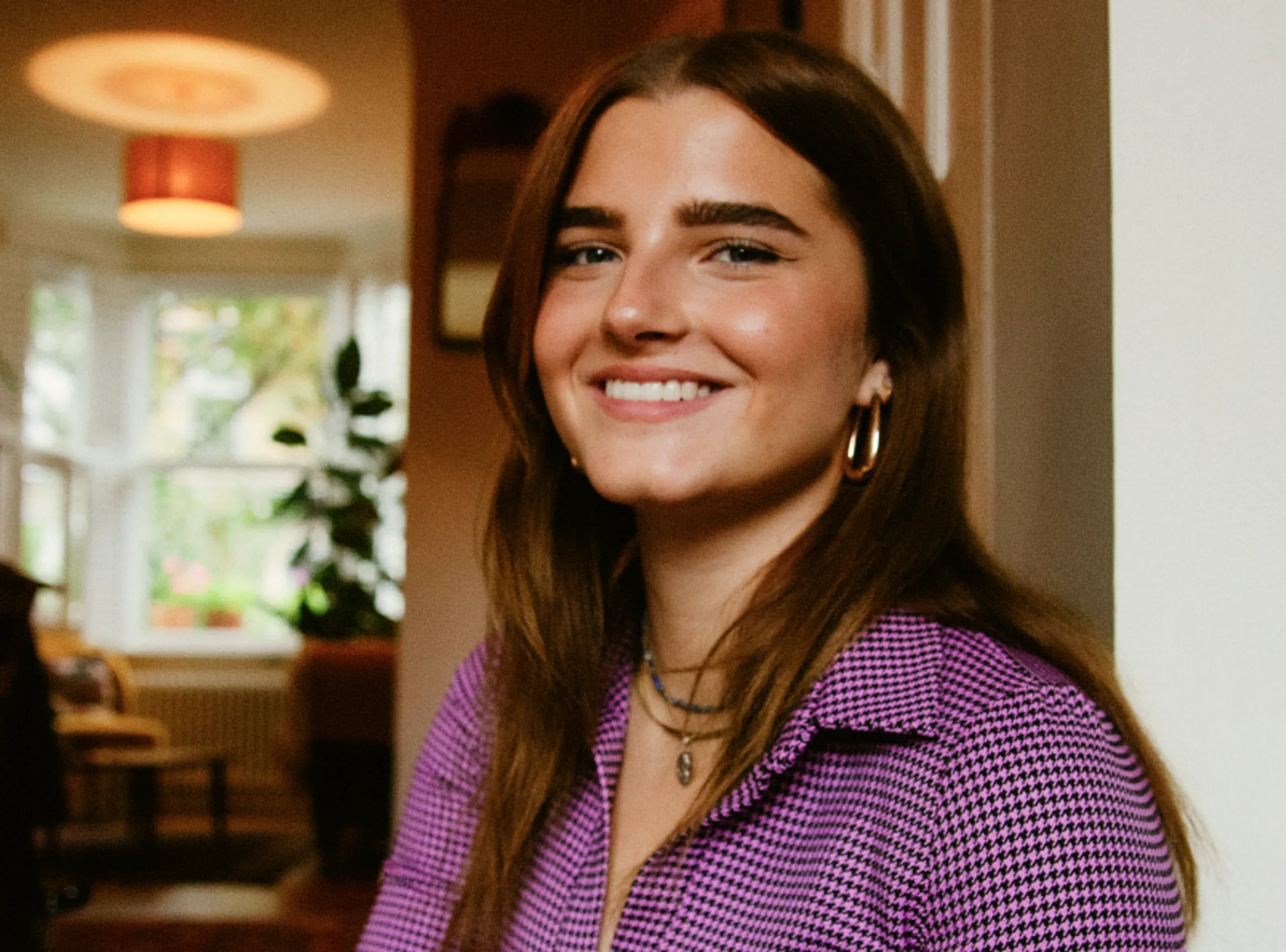 A young woman with long brown hair and hoop earrings smiles indoors, wearing a purple patterned top. A softly lit room with plants and a window is visible in the background.