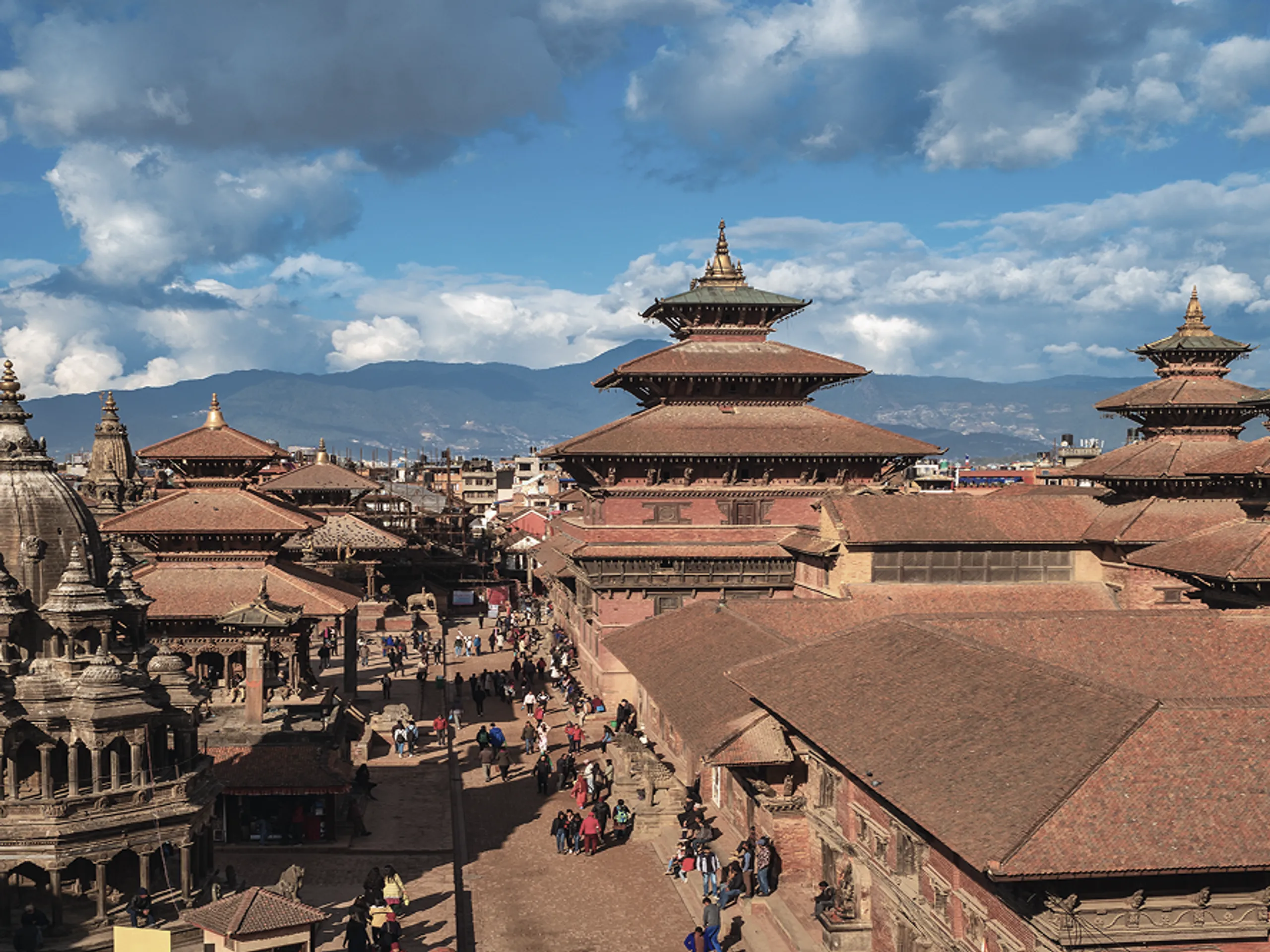 Aerial view of Patan Durbar Square in Nepal, with historic pagoda-style temples, people walking below, and mountains in the background under a partly cloudy sky.