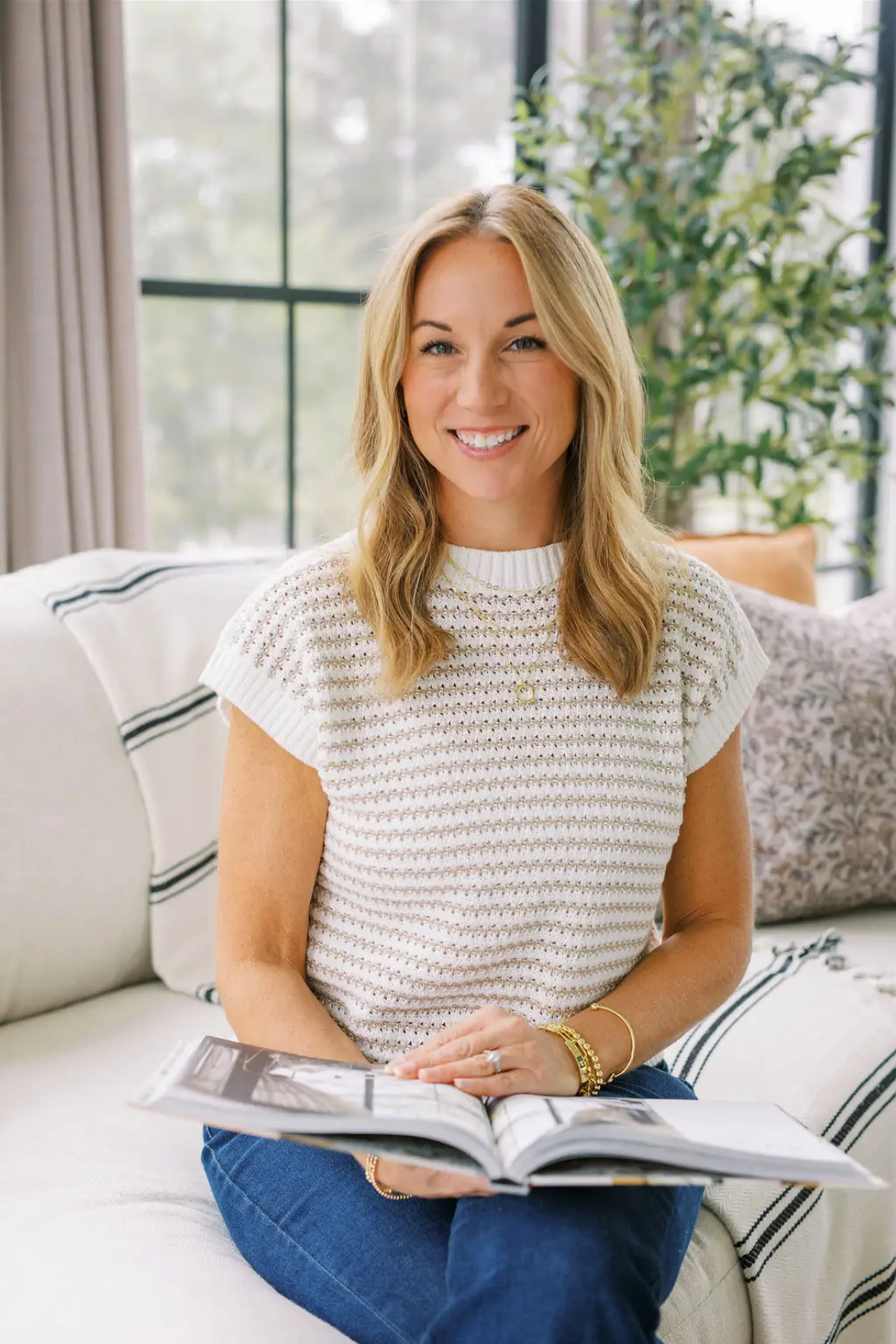 A woman with blonde hair sits on a white couch, smiling while holding an open book. She wears a white knit top and blue jeans, with a plant and large window in the background.