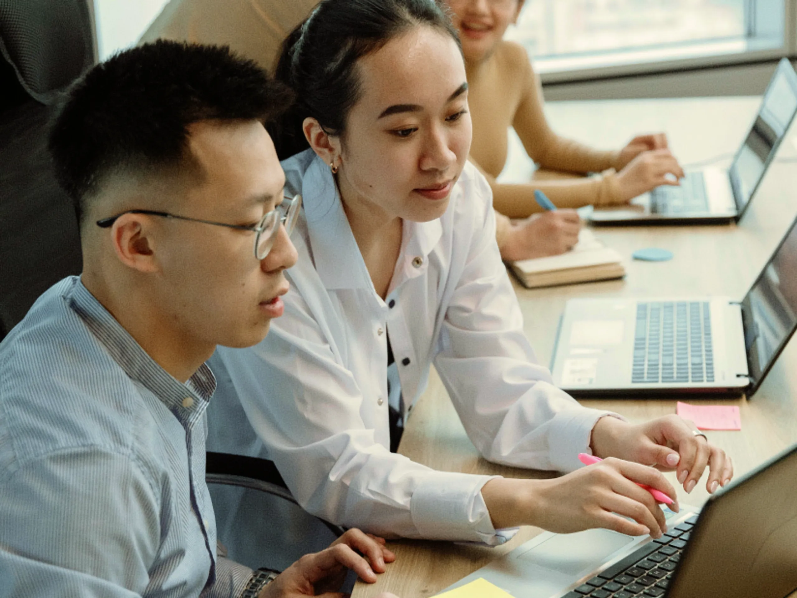 Two people collaborate on a laptop at a desk with another person working nearby. Sticky notes and notebooks are visible, suggesting a team meeting or group project in a modern office setting.
