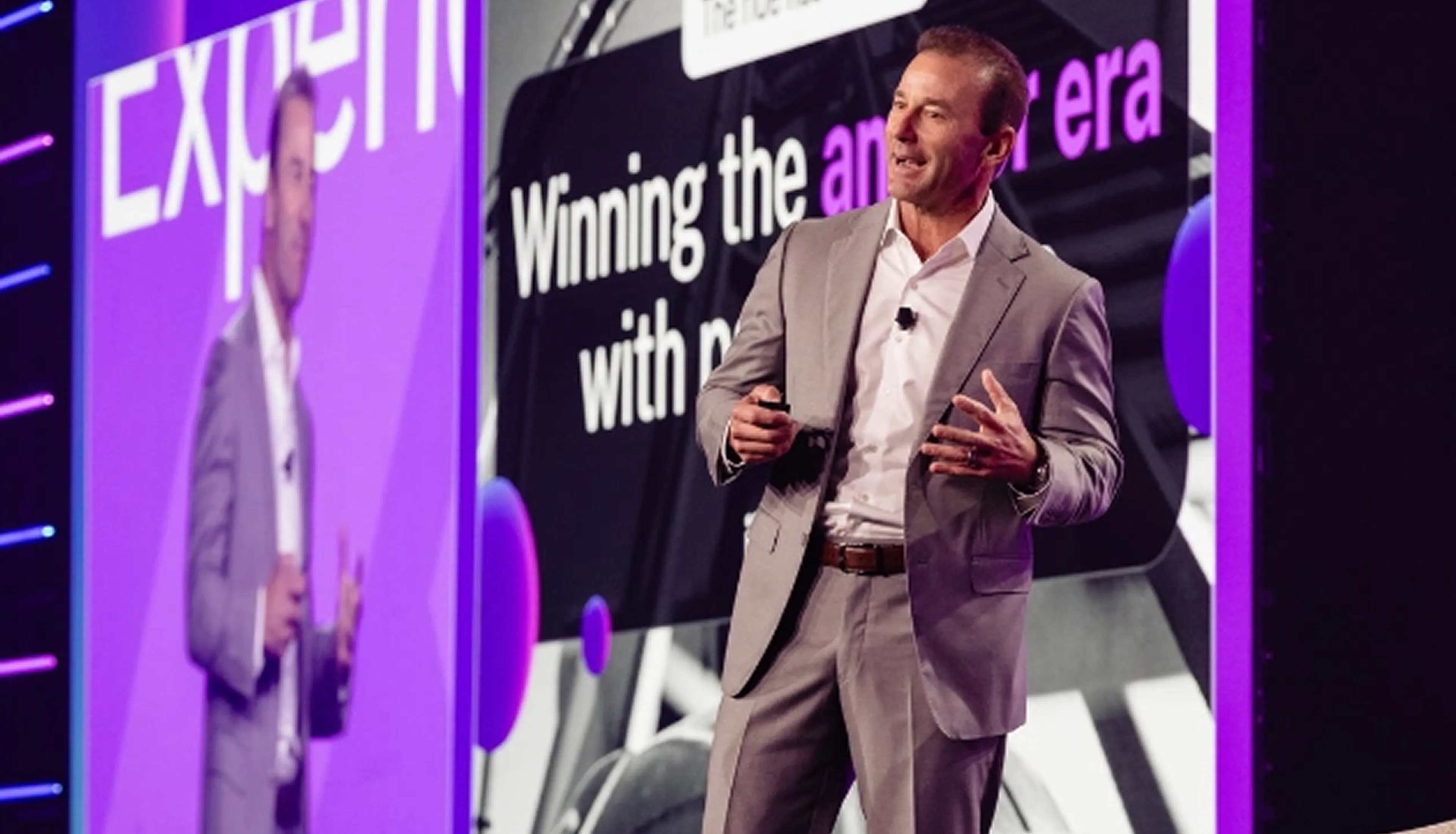 A man in a gray suit speaks on stage in front of a large screen displaying the words Winning the ... era with p.... Purple lighting highlights the scene.