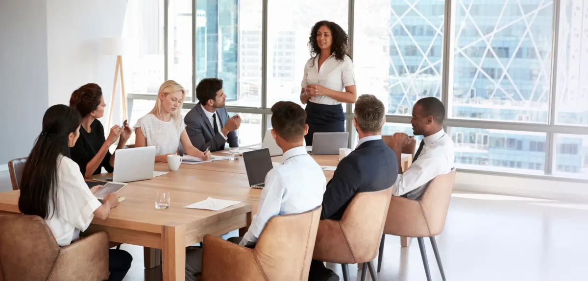 Business meeting in a modern office with eight professionals seated around a wooden table, one woman standing and presenting.
