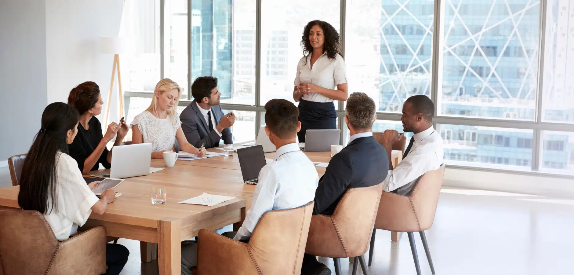 Business meeting in a modern office with eight professionals seated around a wooden table, one woman standing and presenting.