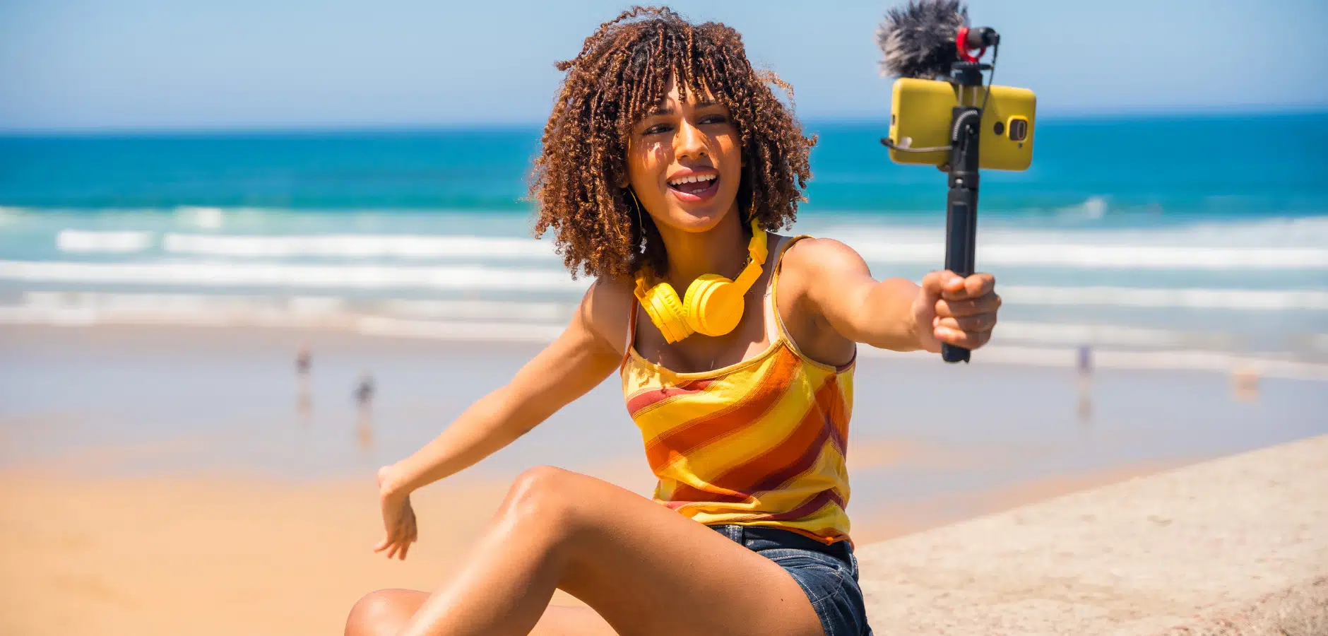 A woman sitting on the beach, smiling while holding a selfie stick to capture her photo against the ocean backdrop.