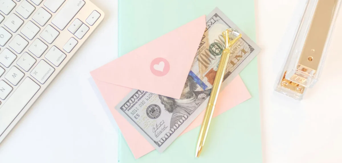 Pink envelopes with a heart, U.S. dollar bills, and a gold pen beside a computer keyboard and stapler.