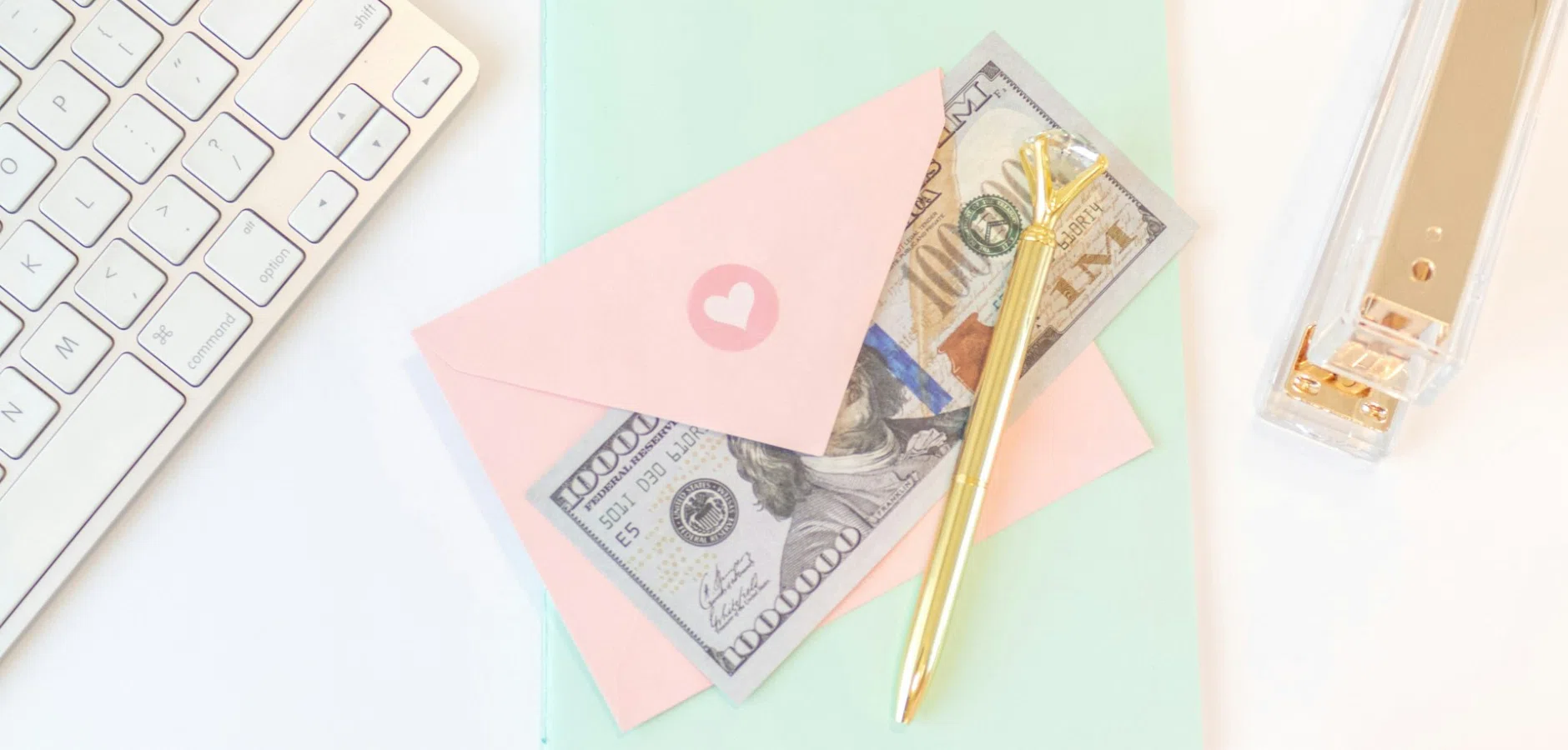 Pink envelopes with a heart, U.S. dollar bills, and a gold pen beside a computer keyboard and stapler.
