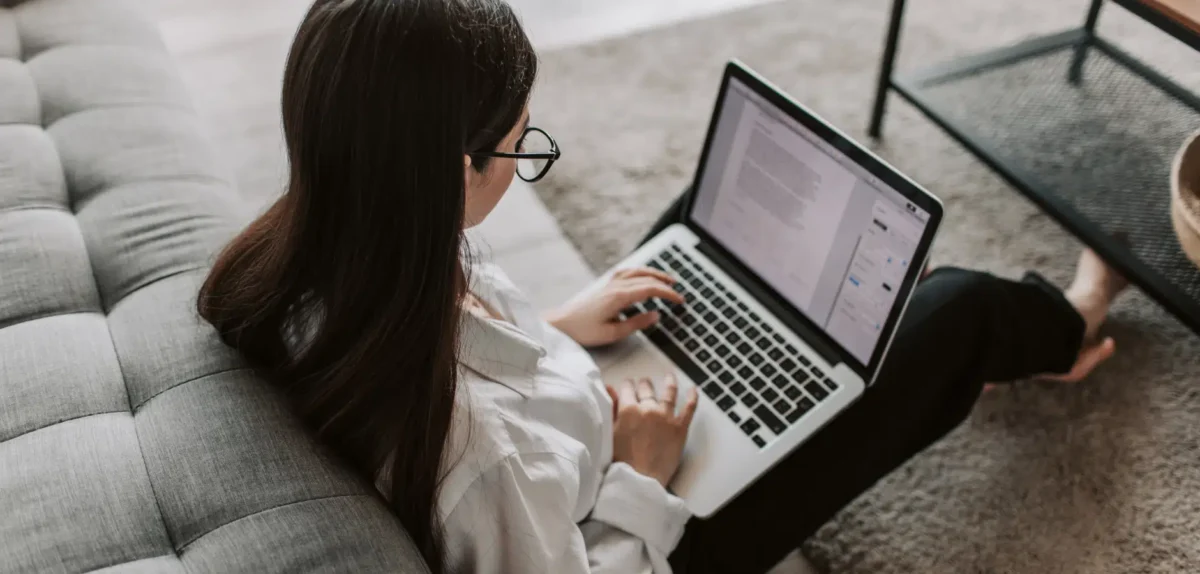 Woman wearing glasses working on a laptop while sitting on the floor next to a gray sofa and a black metal table.