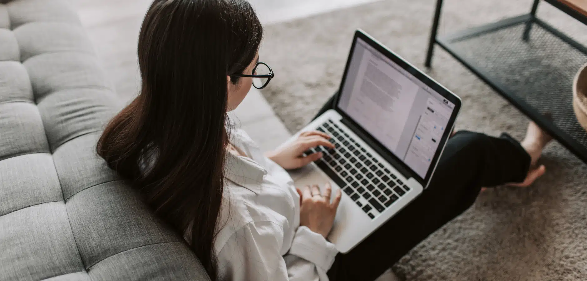 Woman wearing glasses working on a laptop while sitting on the floor next to a gray sofa and a black metal table.