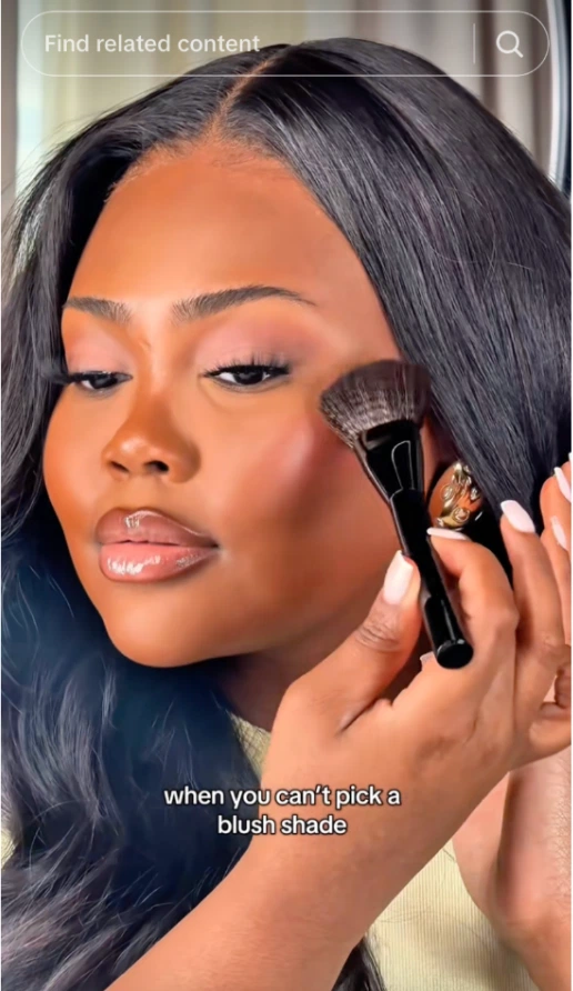 A woman holds a makeup brush near her cheek, contemplating which blush shade to choose, surrounded by soft lighting and elegance.