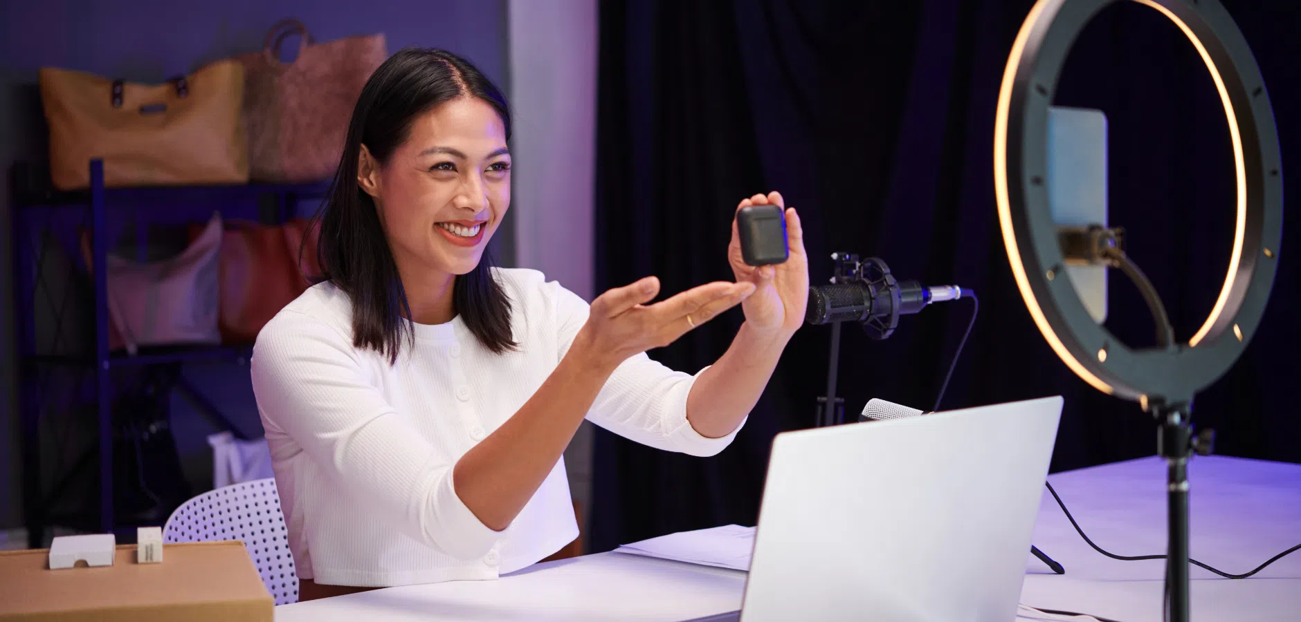 Woman presenting a small black tech gadget while recording video with a ring light, laptop, and microphone in a room with bags on shelves.