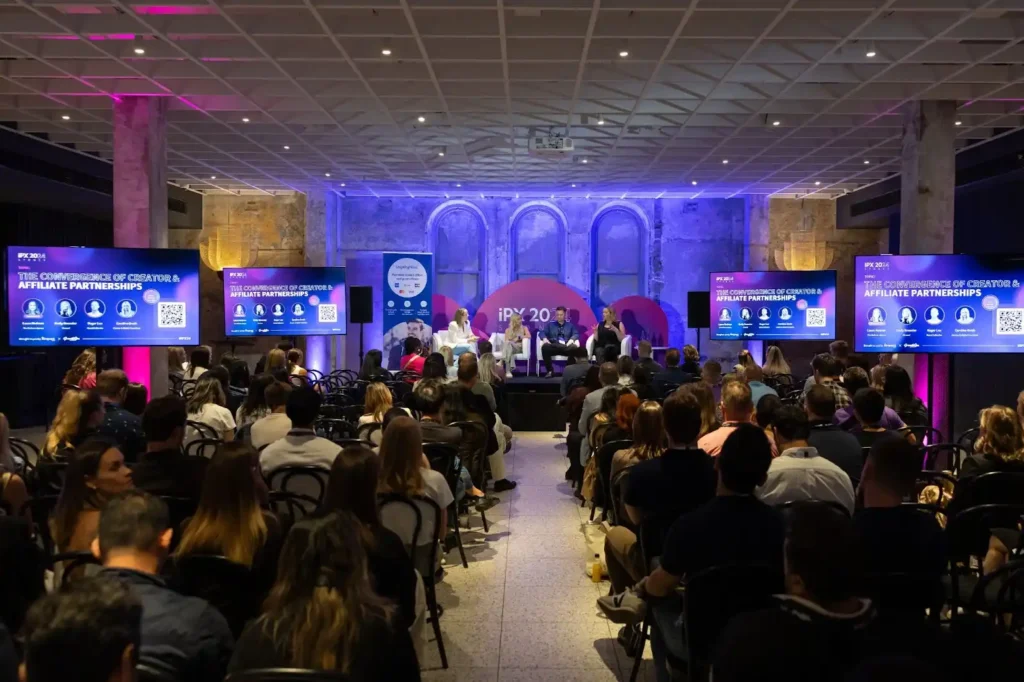 A large crowd attentively watches a presentation by Emily Brewster in a spacious room filled with engaged attendees.