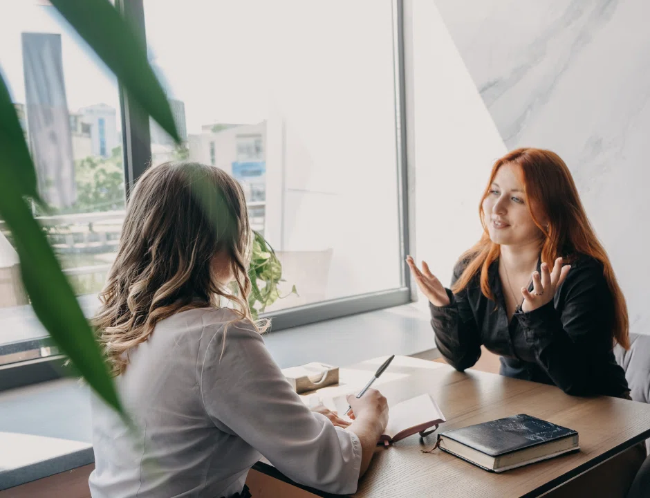 Two women engaged in conversation in a professional office environment, discussing work-related matters.