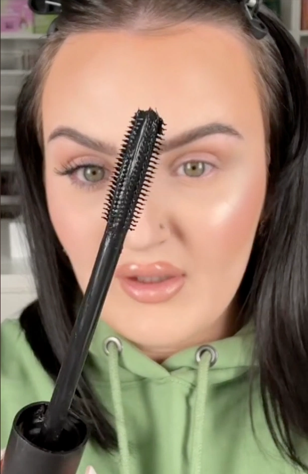 A woman named Mikayla Nogueria holds a mascara brush, preparing to enhance her eyelashes with makeup.