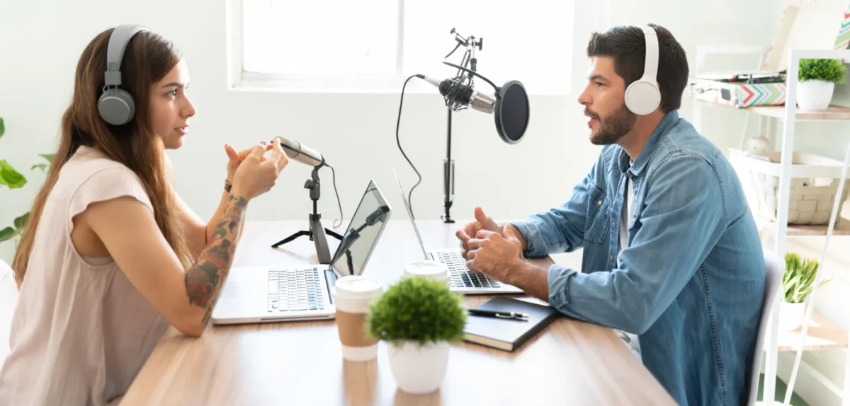 A man and woman wearing headphones sit at a table, engaged in a podcast discussion.