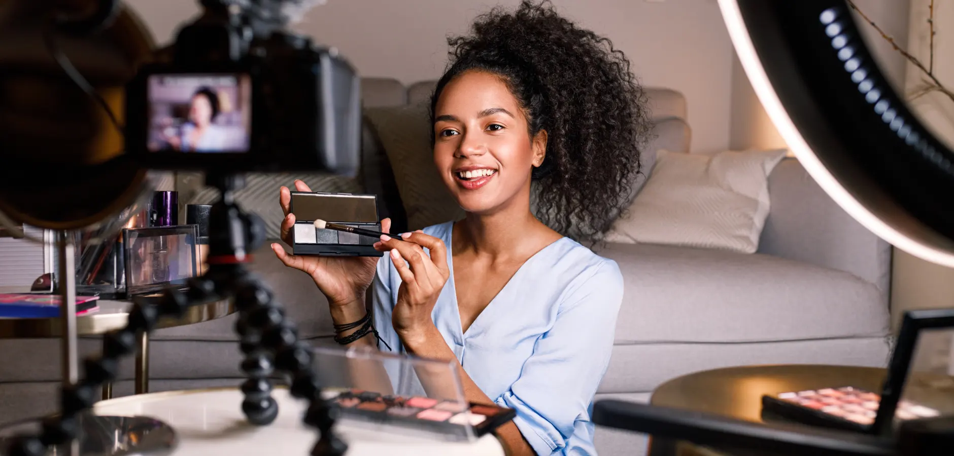 A woman holding a camera in one hand and various makeup products in the other, showcasing her dual passion for photography and beauty.