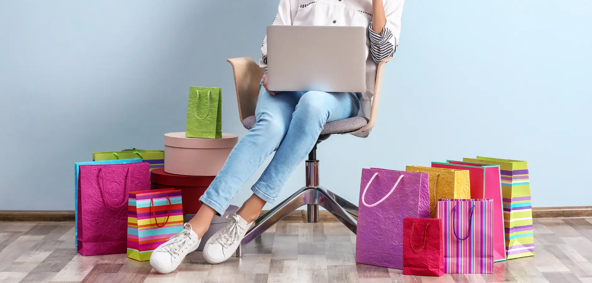 A woman seated on a chair, using a laptop, surrounded by shopping bags, indicating a day of online shopping.