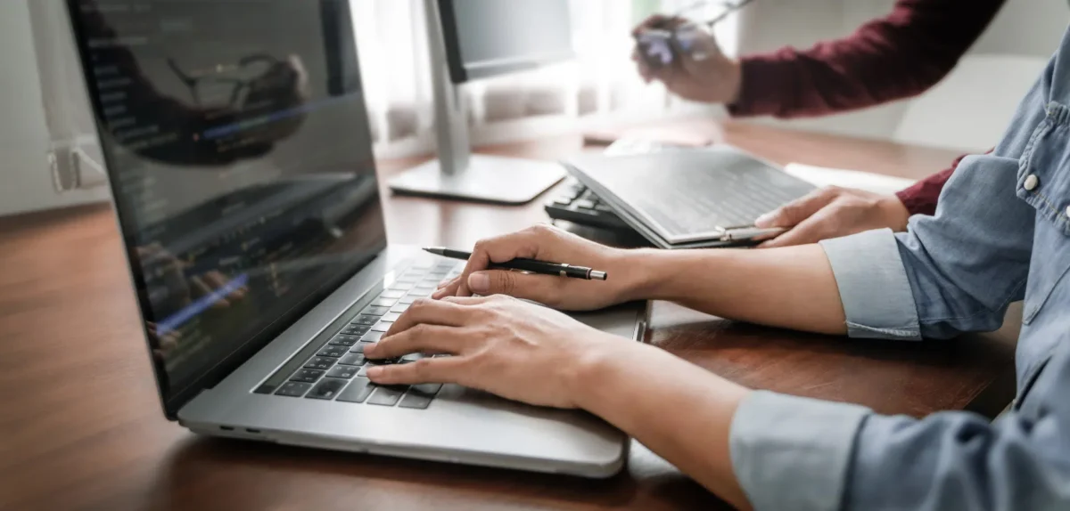 A man focused on his laptop while another individual observes him attentively in a collaborative workspace.