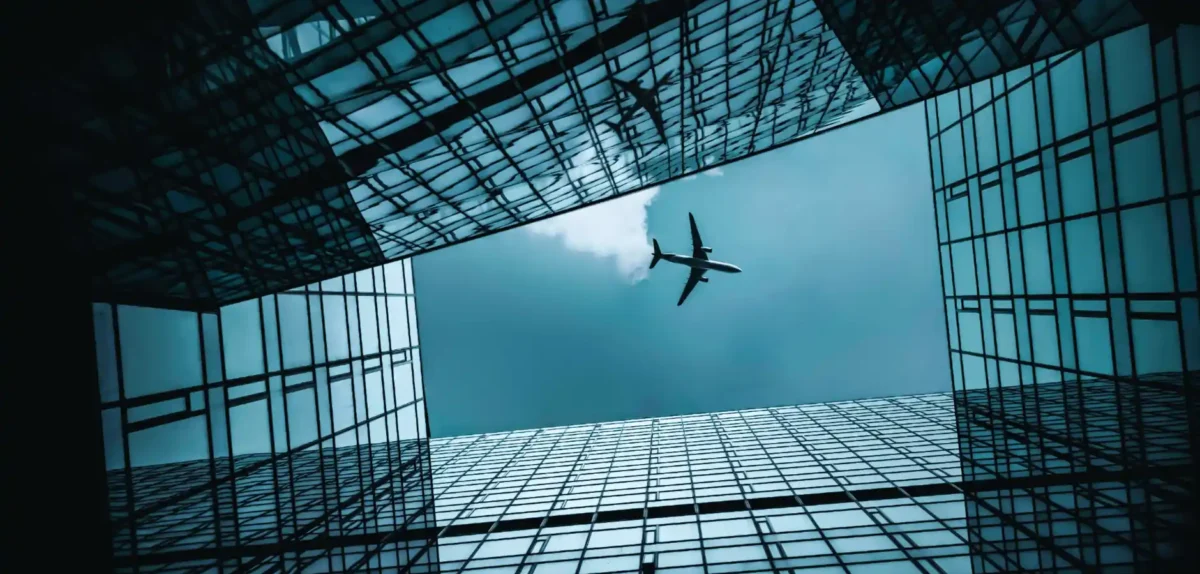 A plane soars through a clear sky above a modern building, showcasing the contrast between aviation and urban architecture.